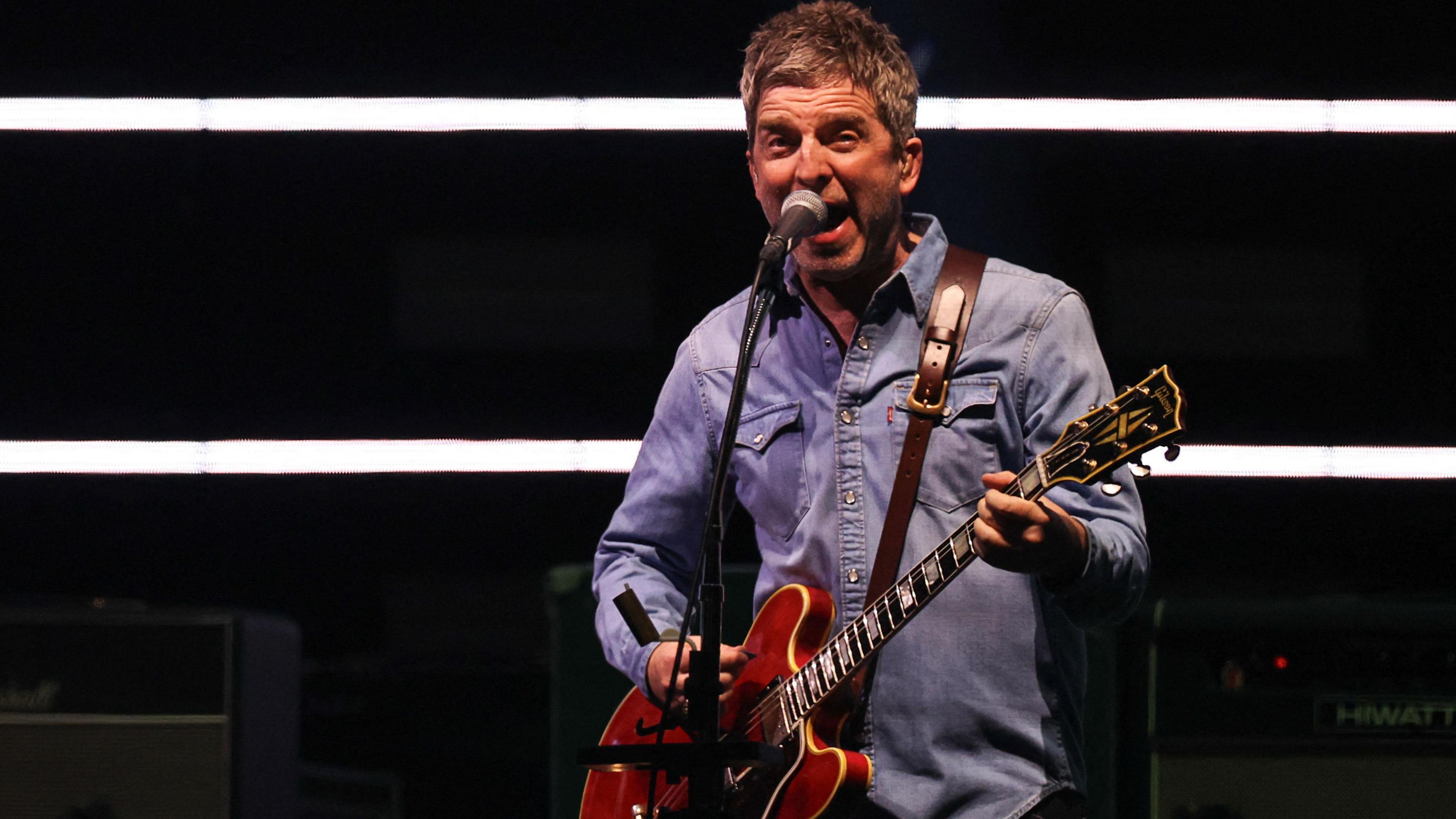 Noel Gallagher in blue denim sings into a microphone and plays a red guitar at the Oasis comeback gig in Cardiff