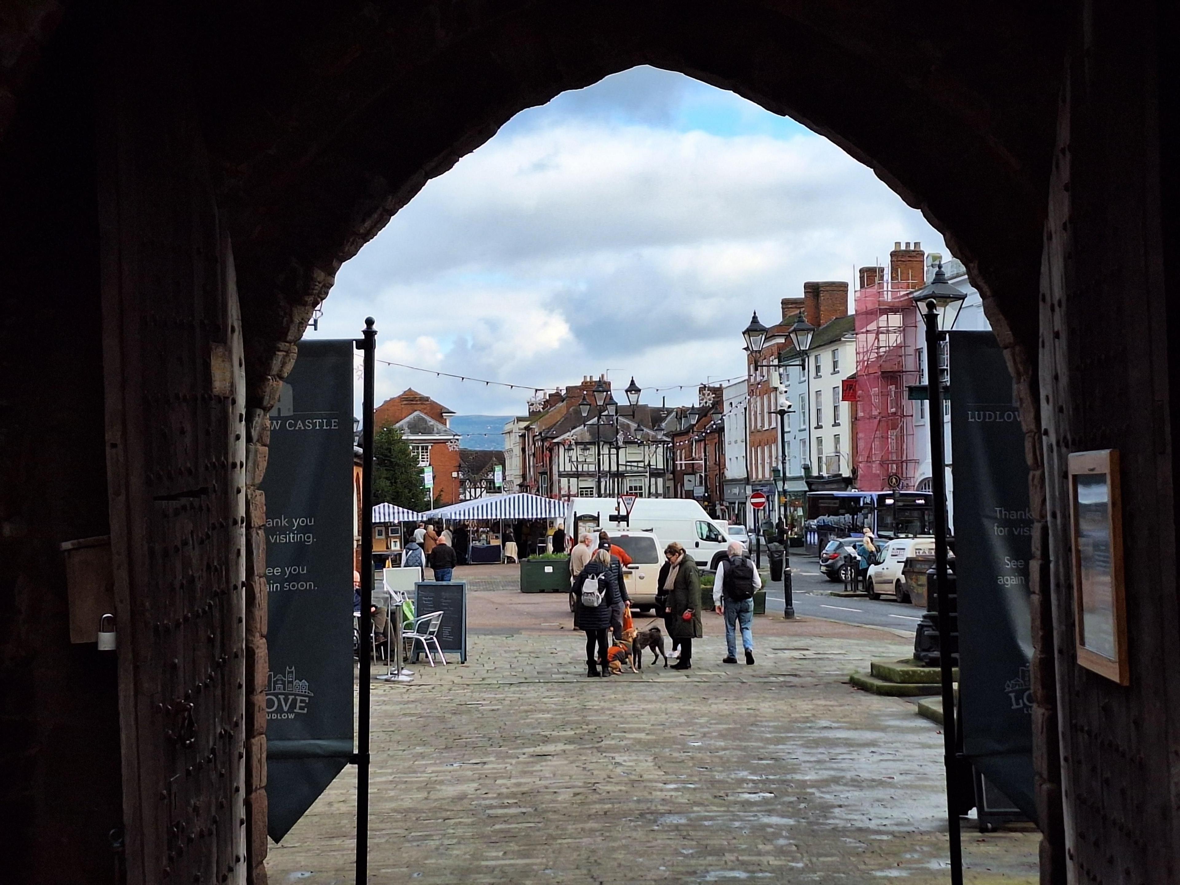 A gateway with a pointed arch silhouettes a shot of a traditional town square with a range of heritage buildings and covered market stalls with blue and white striped canopies. 