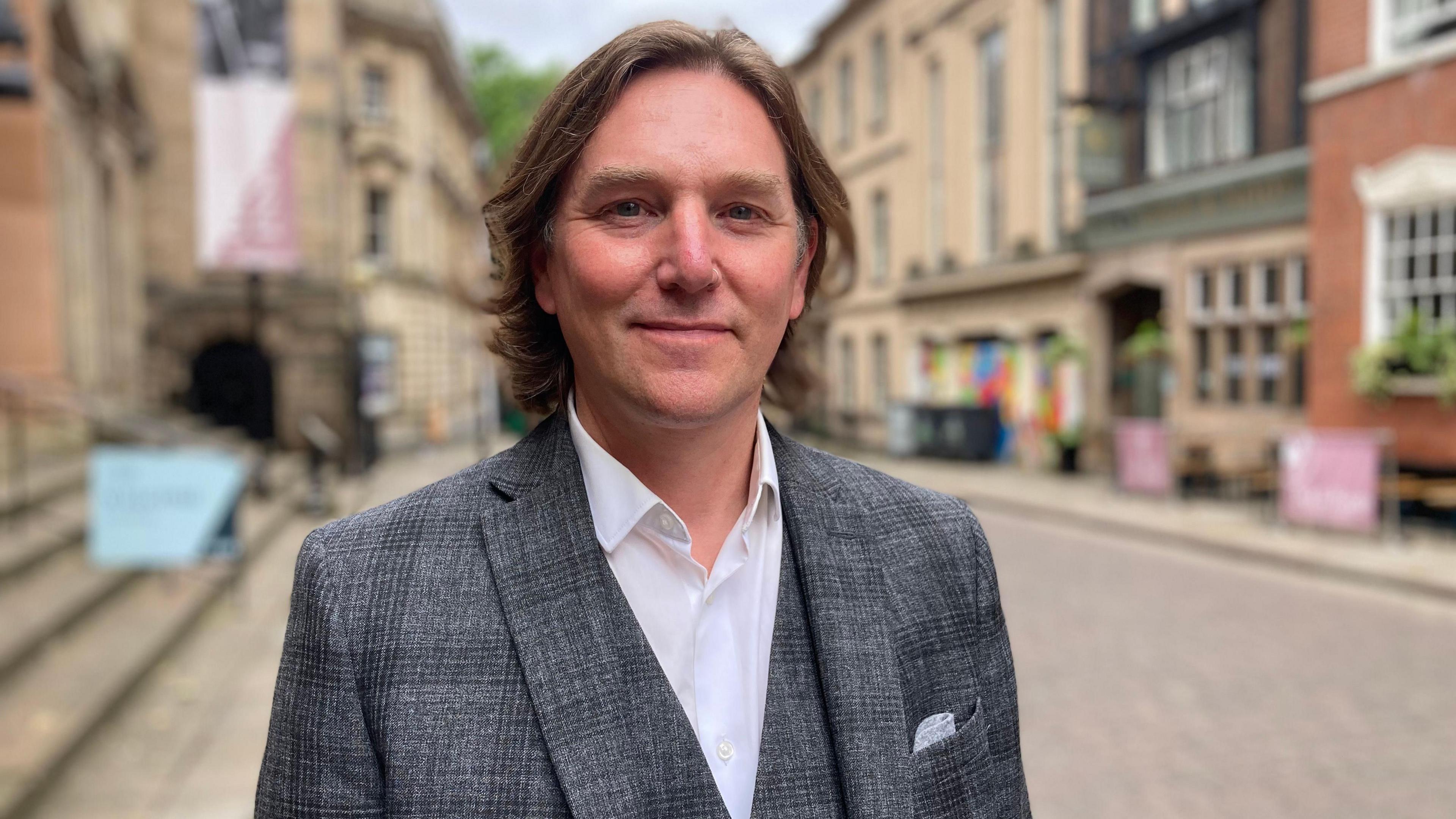 Andrew Napier is pictured wearing a grey checked suit and waistcoat with open collared white shirt. He has shoulder-length brown hair