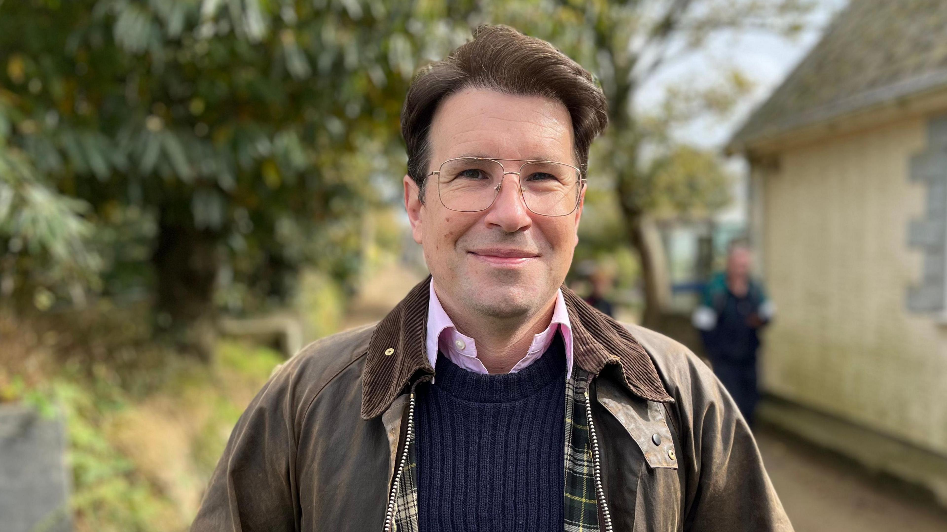 A man with a brown jacket and blue jumper wearing glasses and smiling looking at the camera. 