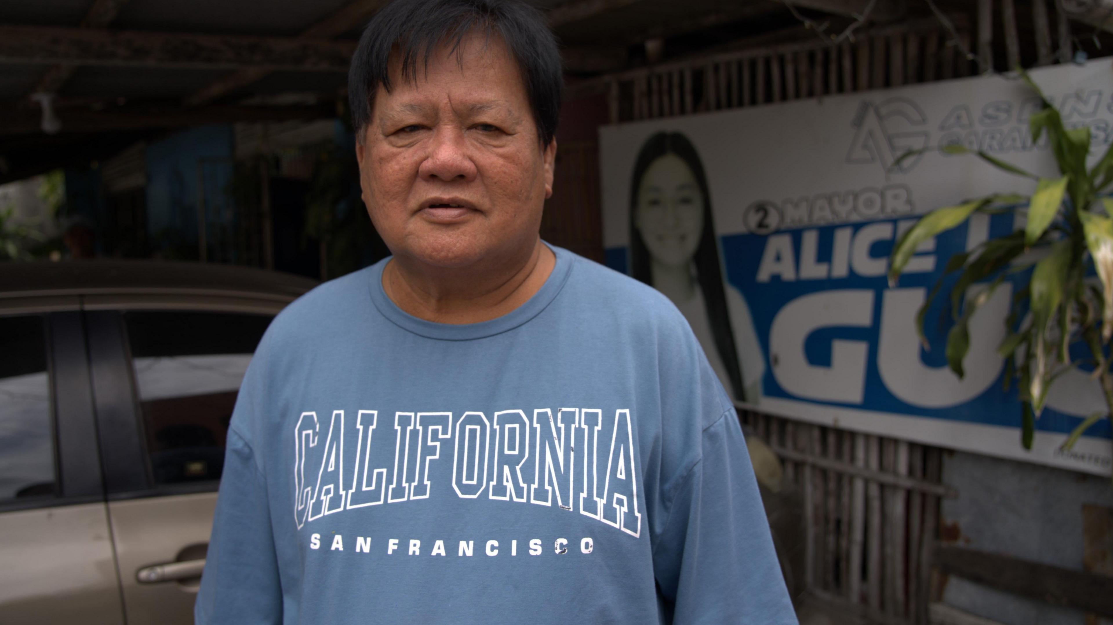 A man dressed in a loose-fitting blue t-shirt with "California, San Francisco" emblazoned across the chest stands in front of a faded campaign poster bearing the words "Mayor Alice Leal Guo"