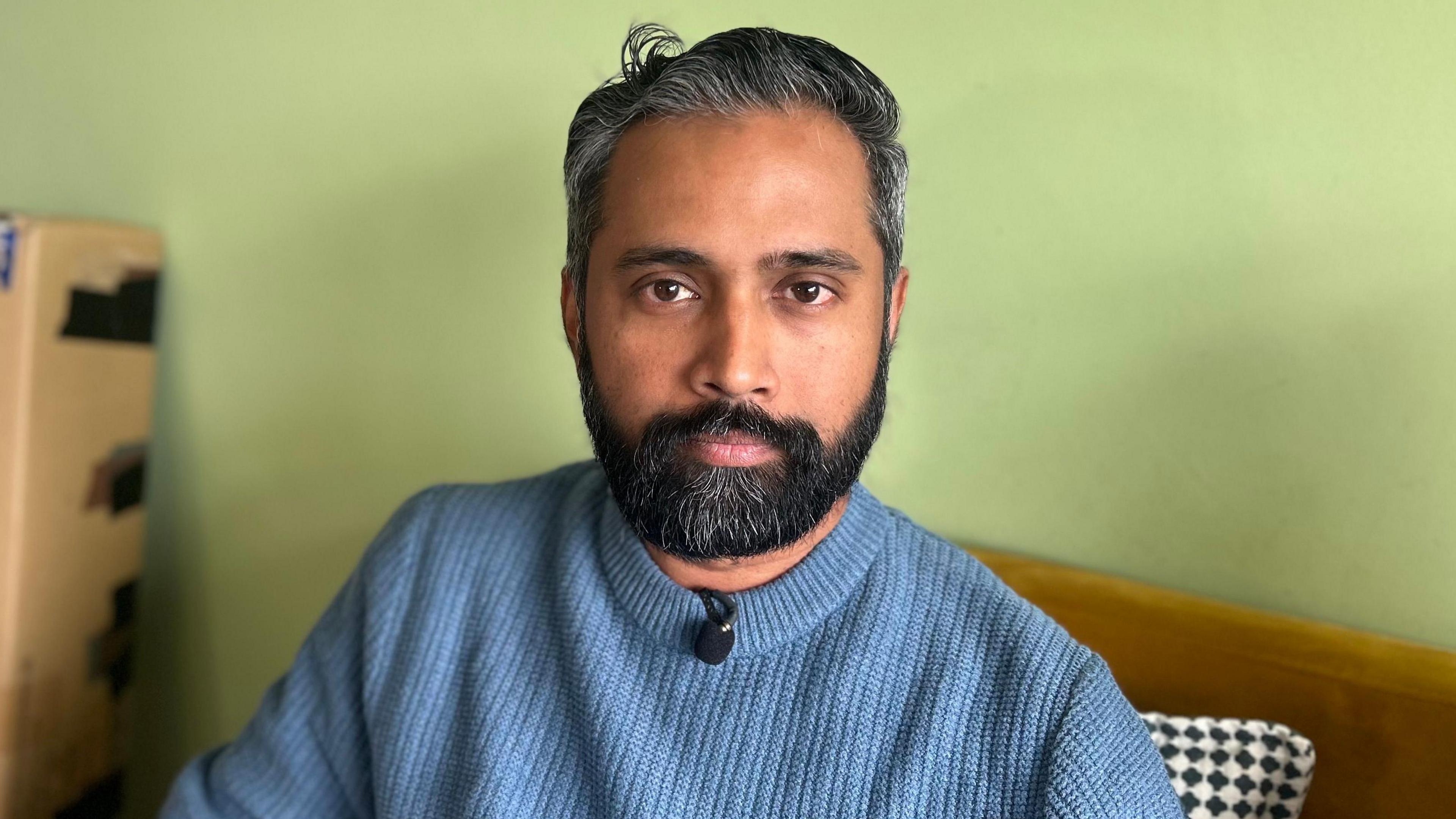 A man, Sree Kadalyil, looks into the camera. He is sitting on an orange sofa that is up against a light green wall. He has short, greying black hair and a black beard that is speckled with grey. He is wearing a light blue jumper. 
