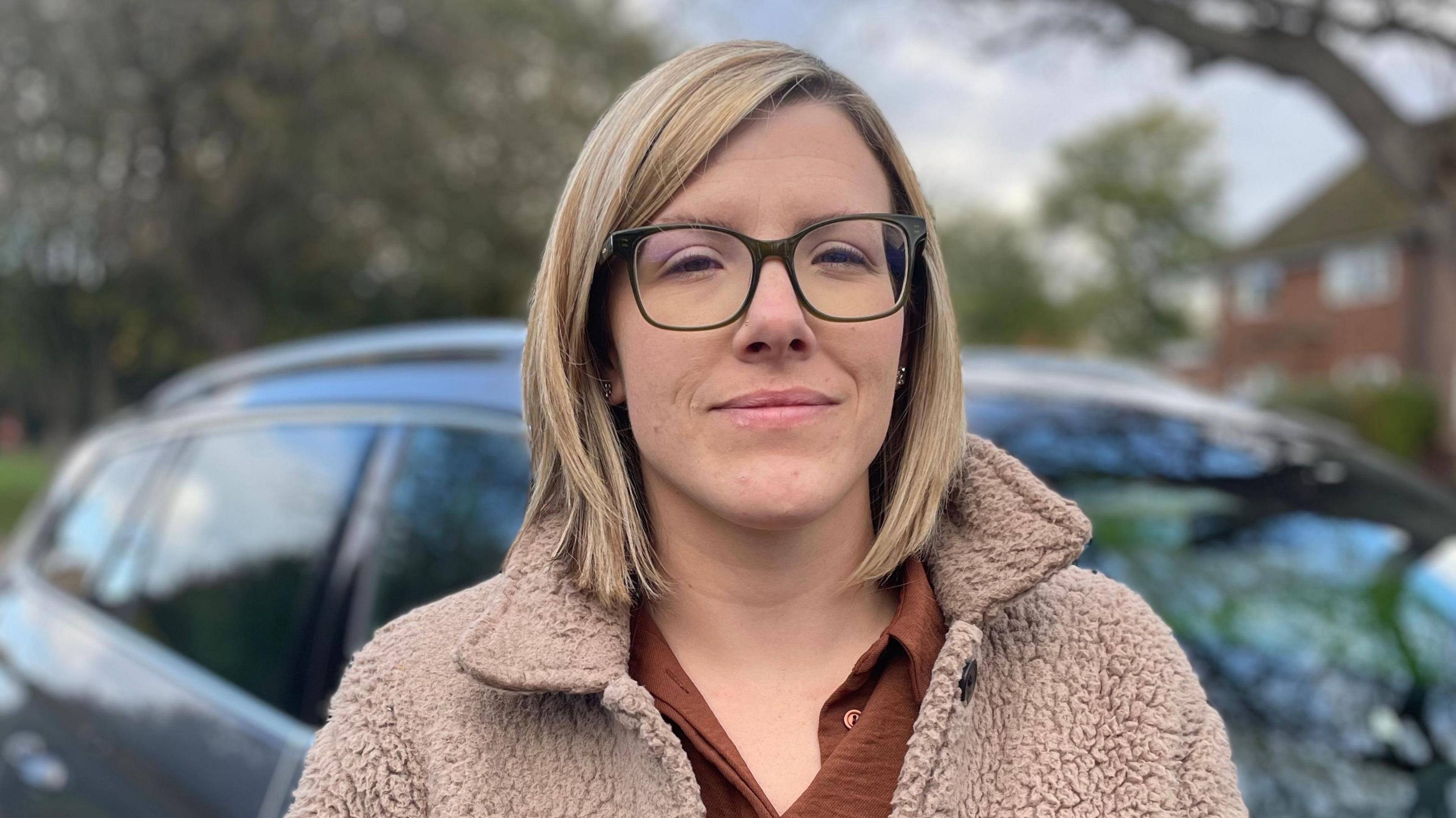 Eve Matthews, looking direct to camera. She is standing outside and in front of a dark-coloured car, parked near some houses. The background is blurred, but one house can be seen to the right of the image, along with some trees. Eve is wearing a cream coat, with the collar turned up, and a rust-coloured top underneath. She has glasses and blond hair cut into a bob. She is smiling weakly. 