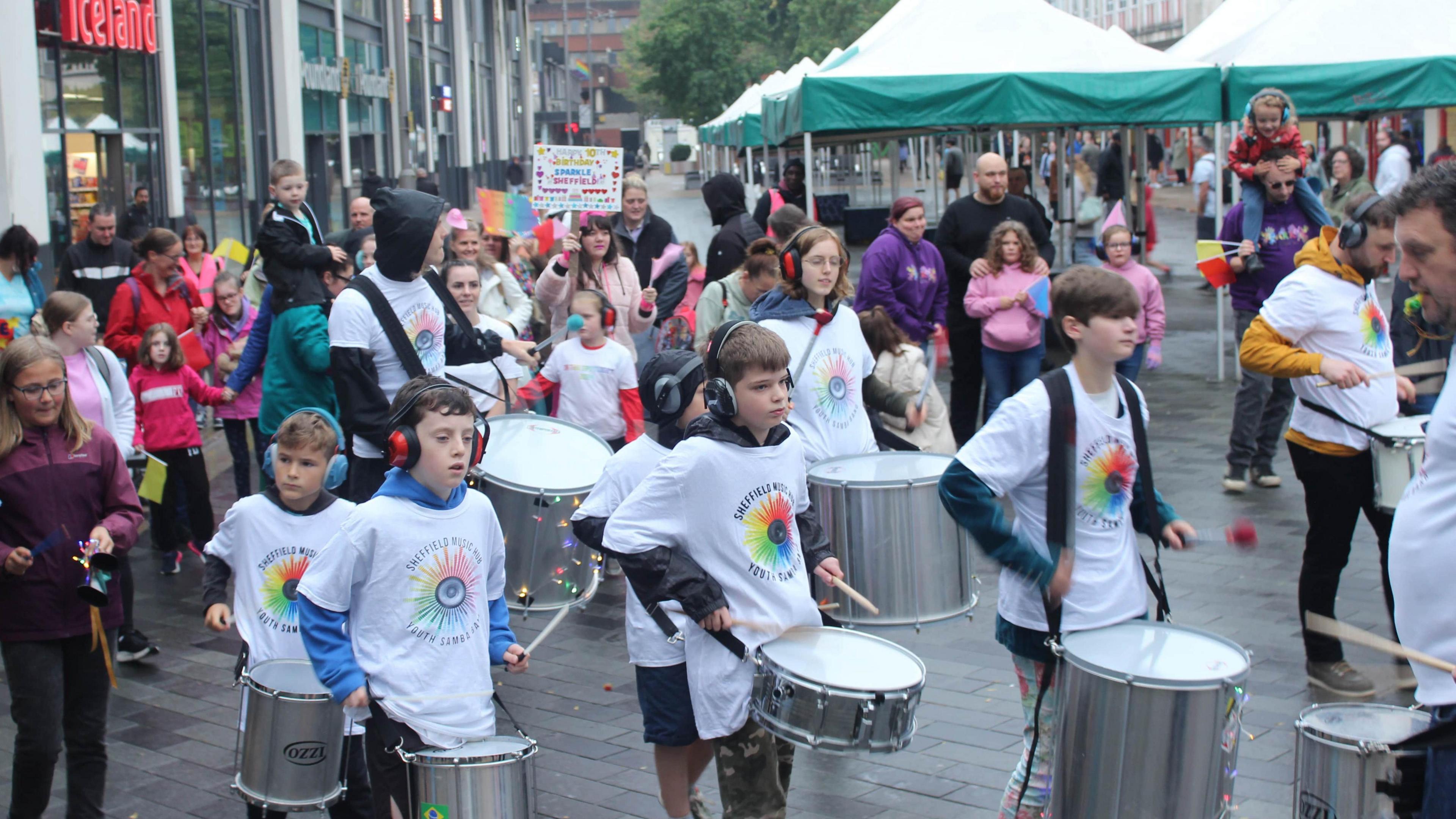 A group of children wearing white t-shirts and headphones play the drums in a busy shopping area 