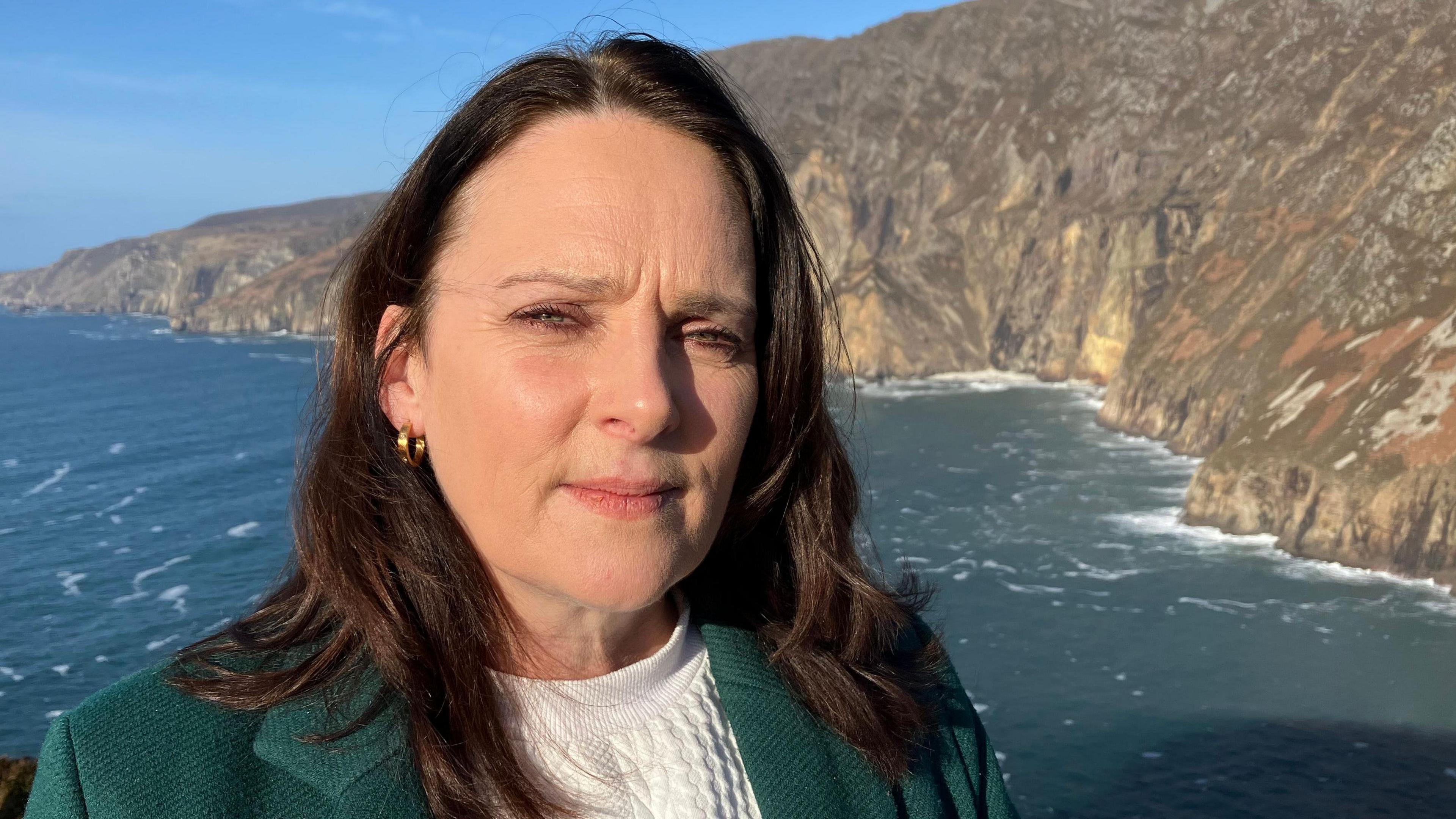 Niamh Kennedy is standing in front of Slieve League. The foot of the cliffs are visible, as is water. She has long, dark hair and is staring into the camera. She is wearing a green jacket with a cream sweater underneath.