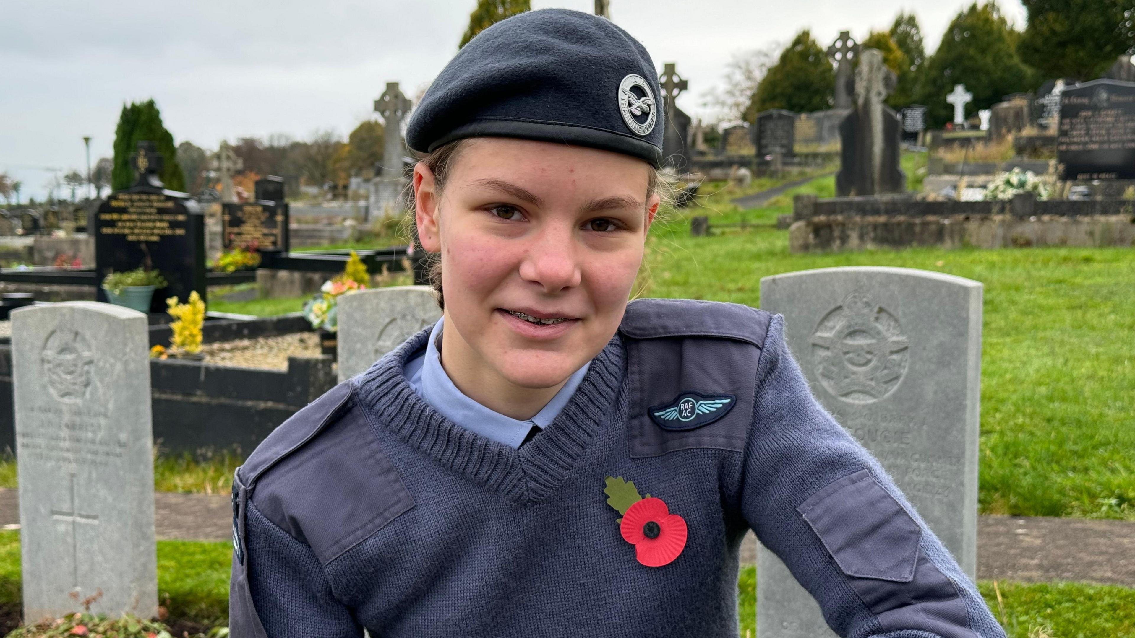 A young female air cadet in blue uniform and beret wearing a poppy. She is kneeling in a cemetery in front of a number of war graves. 