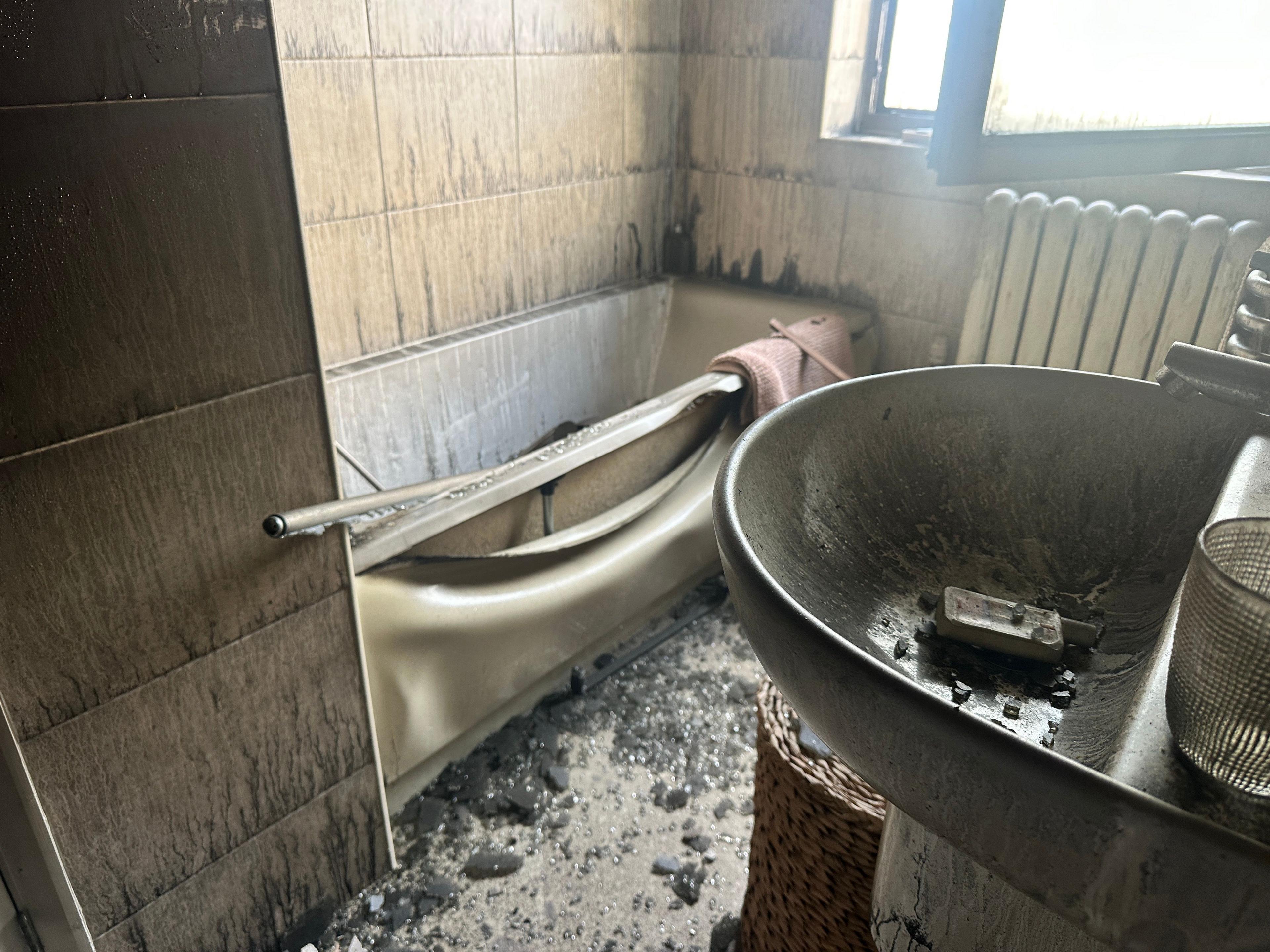 Smoke damage in the bathroom. There is charred remains in the sink and the bath. The walls are blackened and there is smoke damage throughout the white bathroom which is now sooty and black. 