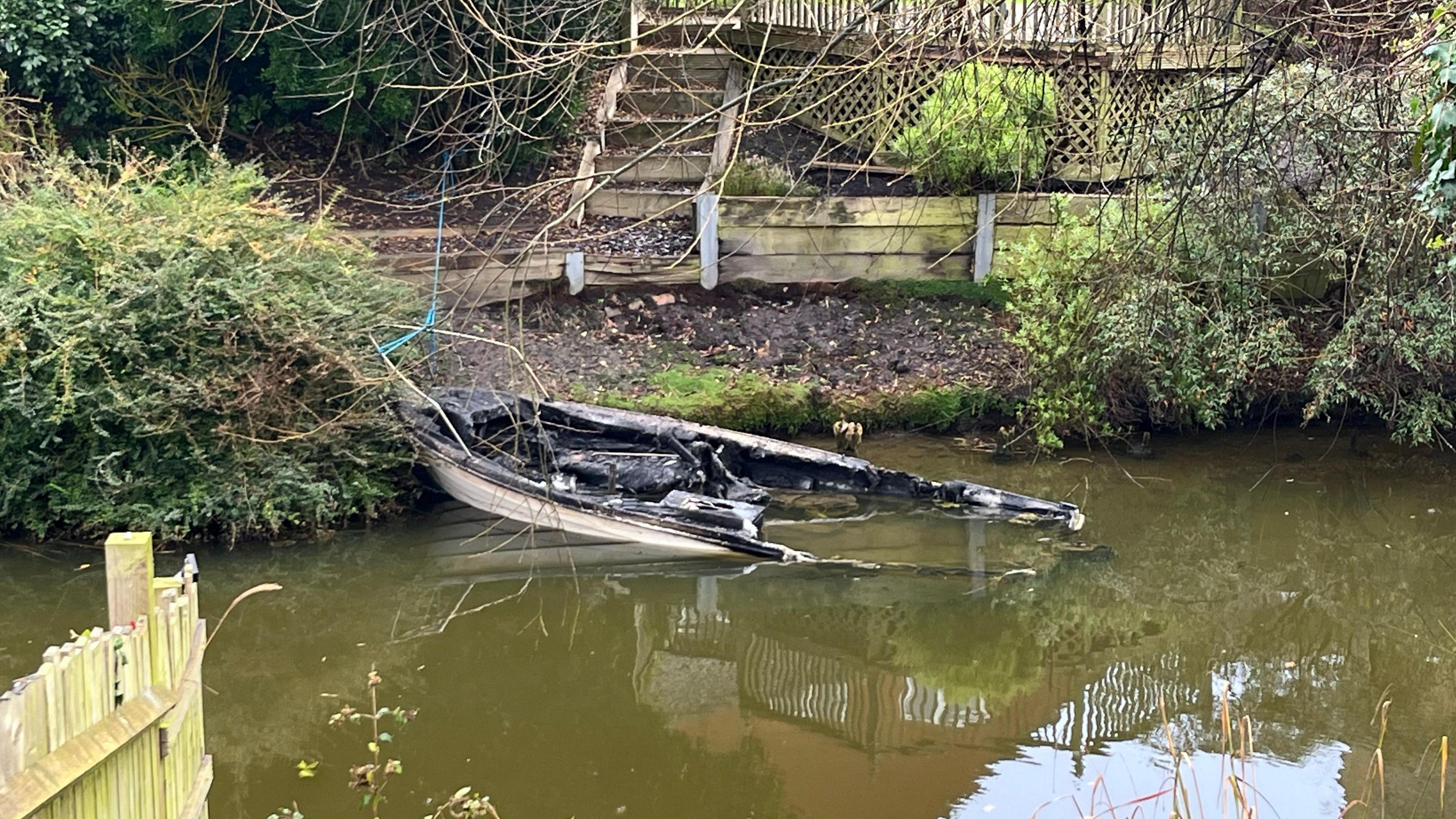 Burnt out hull of a cabin cruiser, about 4.5m long (15ft) sitting in the river near the bankside. The bank is steep, only small, around 1m high leading up to some wooden fencing.