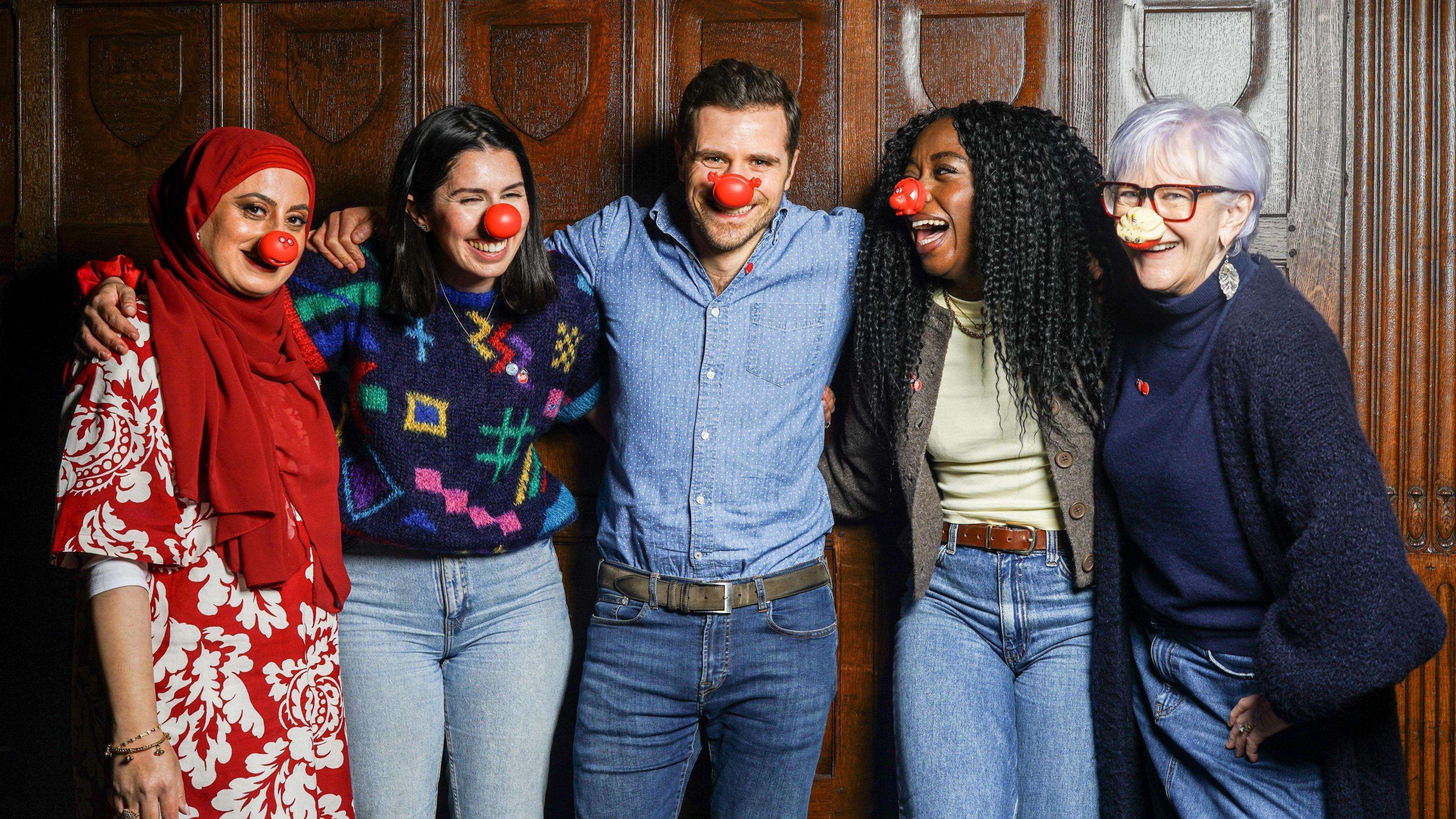 Four women and one man, all wearing red noses, standing against a wood panelled wall with their arms round each other and smiling.