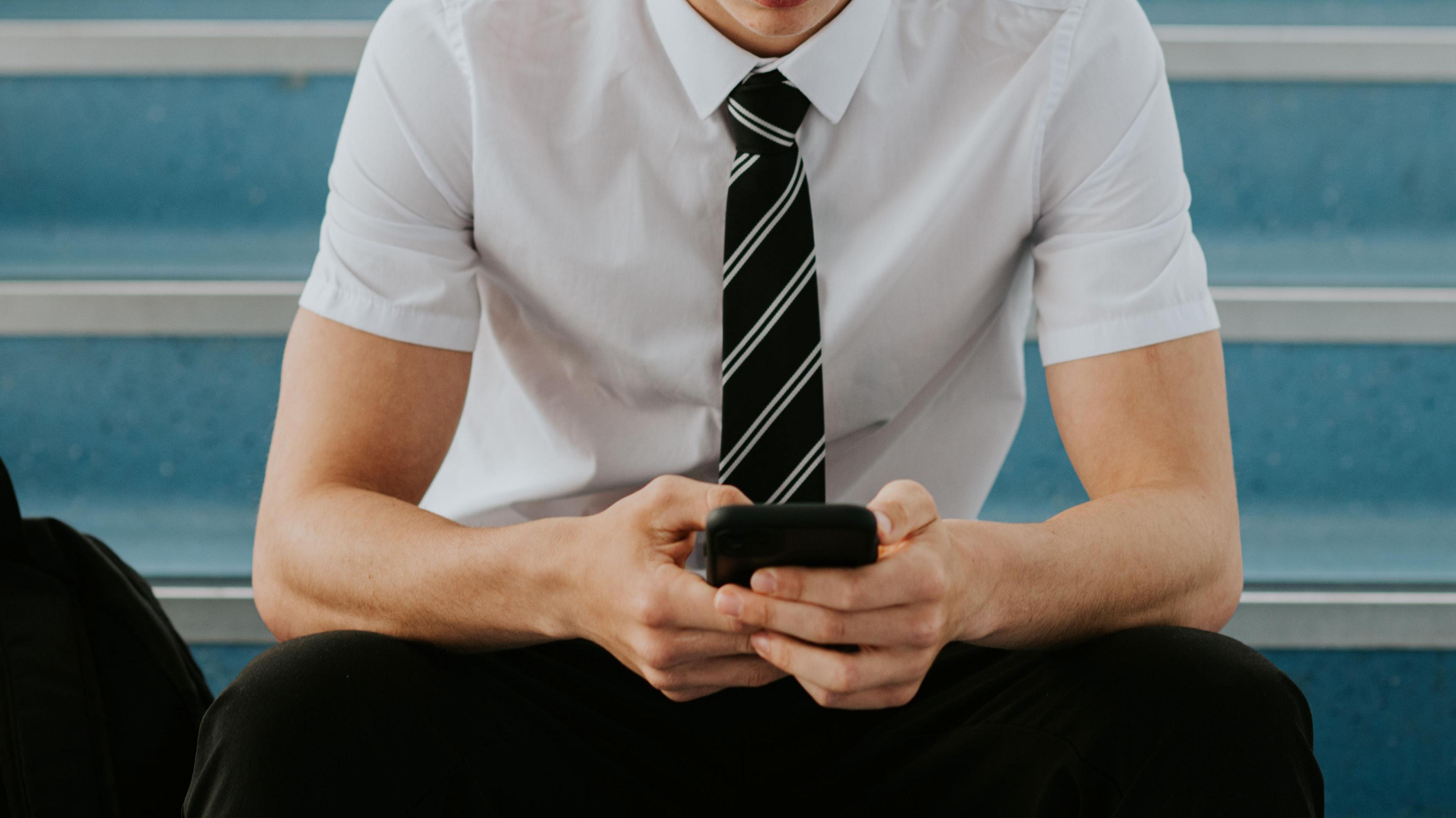 A close-up of a teenage boy as he sits on some steps. He is clutching a smartphone, looking down at it. His face is not visible in the cropped photo.