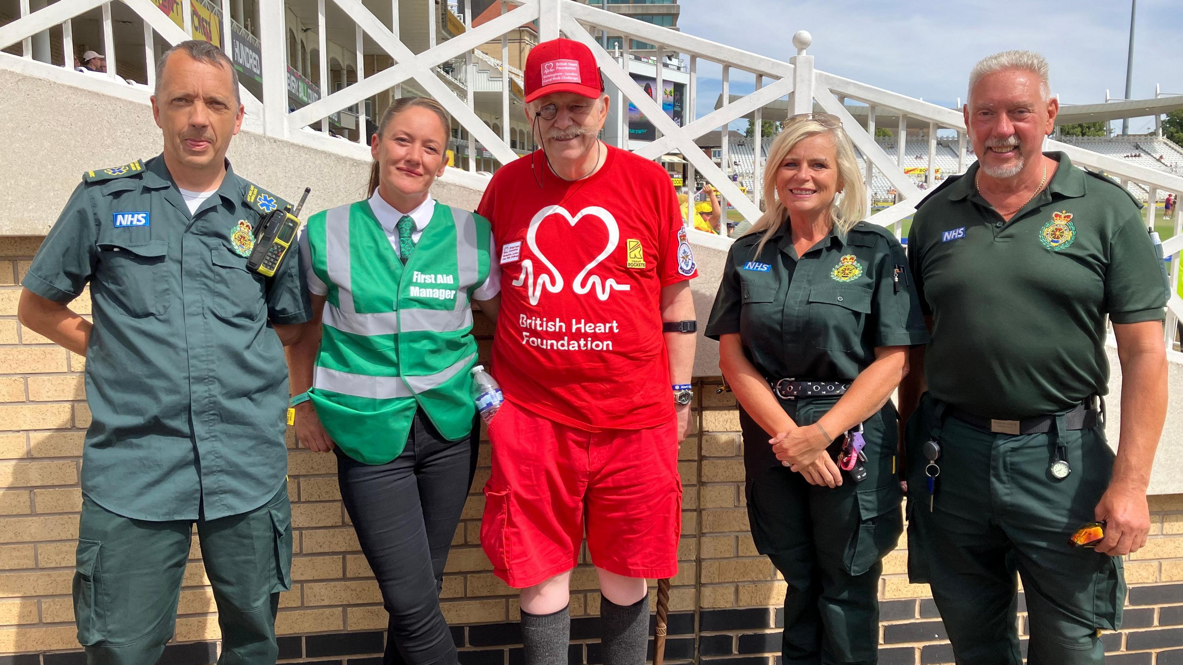 David stands in the middle of a ground of first aiders and paramedics. To David's right is Hayley, wearing a green high-visibility jacket and a green tie. Three paramedics in green uniforms are also standing with David. They all stand in front of one of the stands at Trent Bridge Cricket Ground.