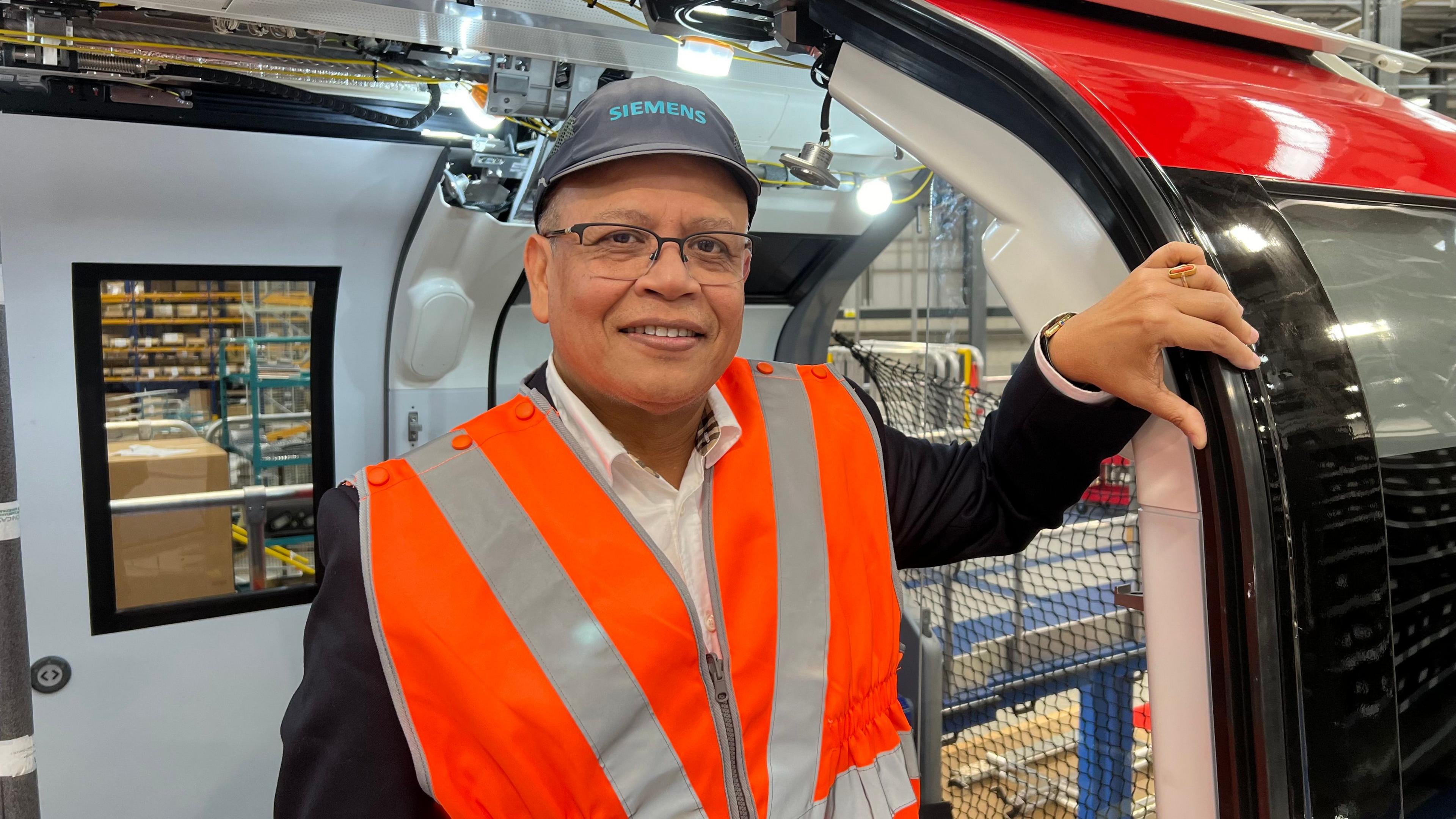 Mr Banerjee wearing an orange high-vis vest and a black cap with Siemens on it. His left hand is holding on to the doorframe of a tube train