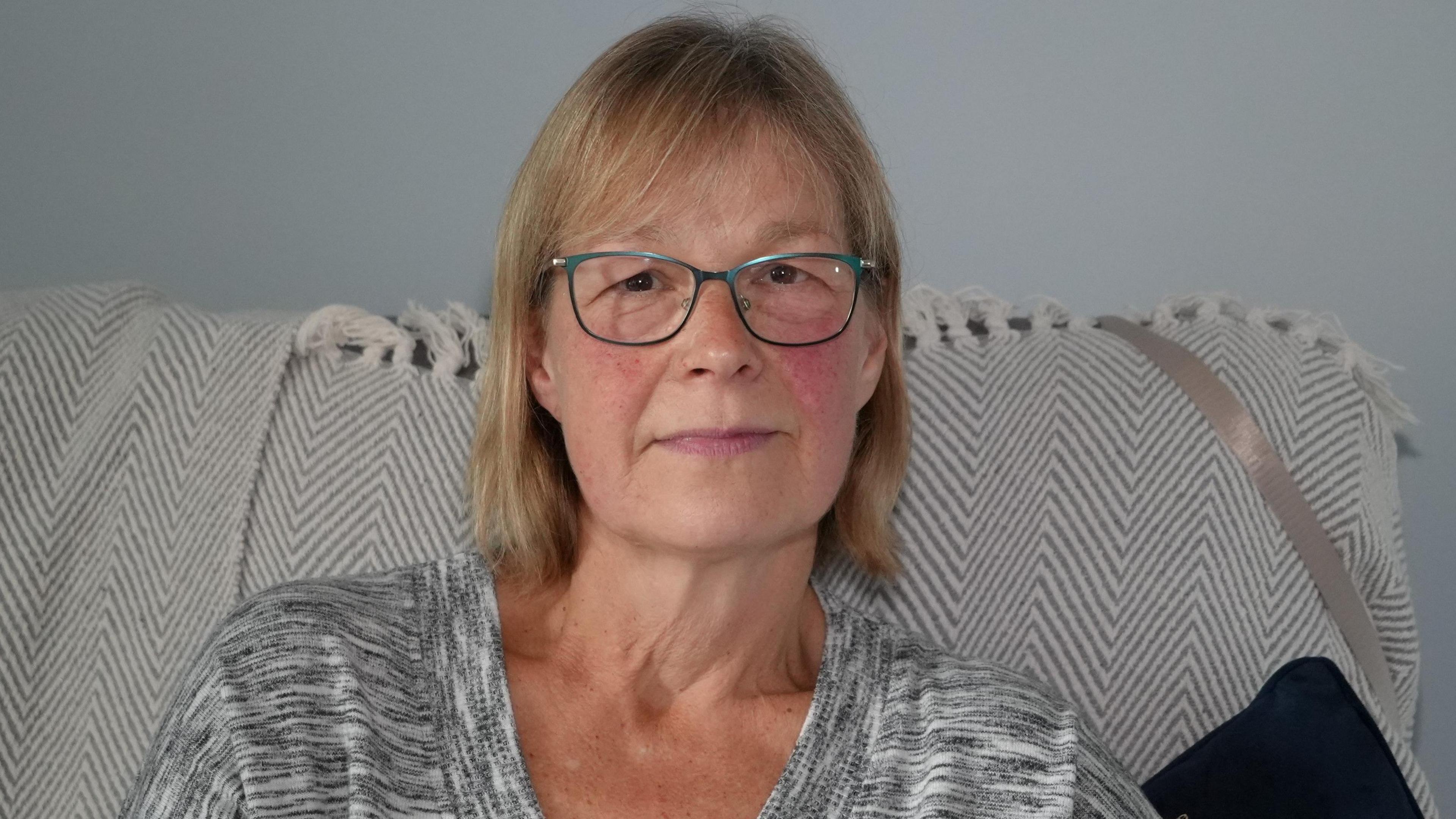 Alison Kingstone-Miles who is sitting on a sofa which has a cream and grey throw on it. She is wearing glasses with blue frames and is looking directly at the camera and smiling. She is wearing a grey and white striped long-sleeve T-shirt.