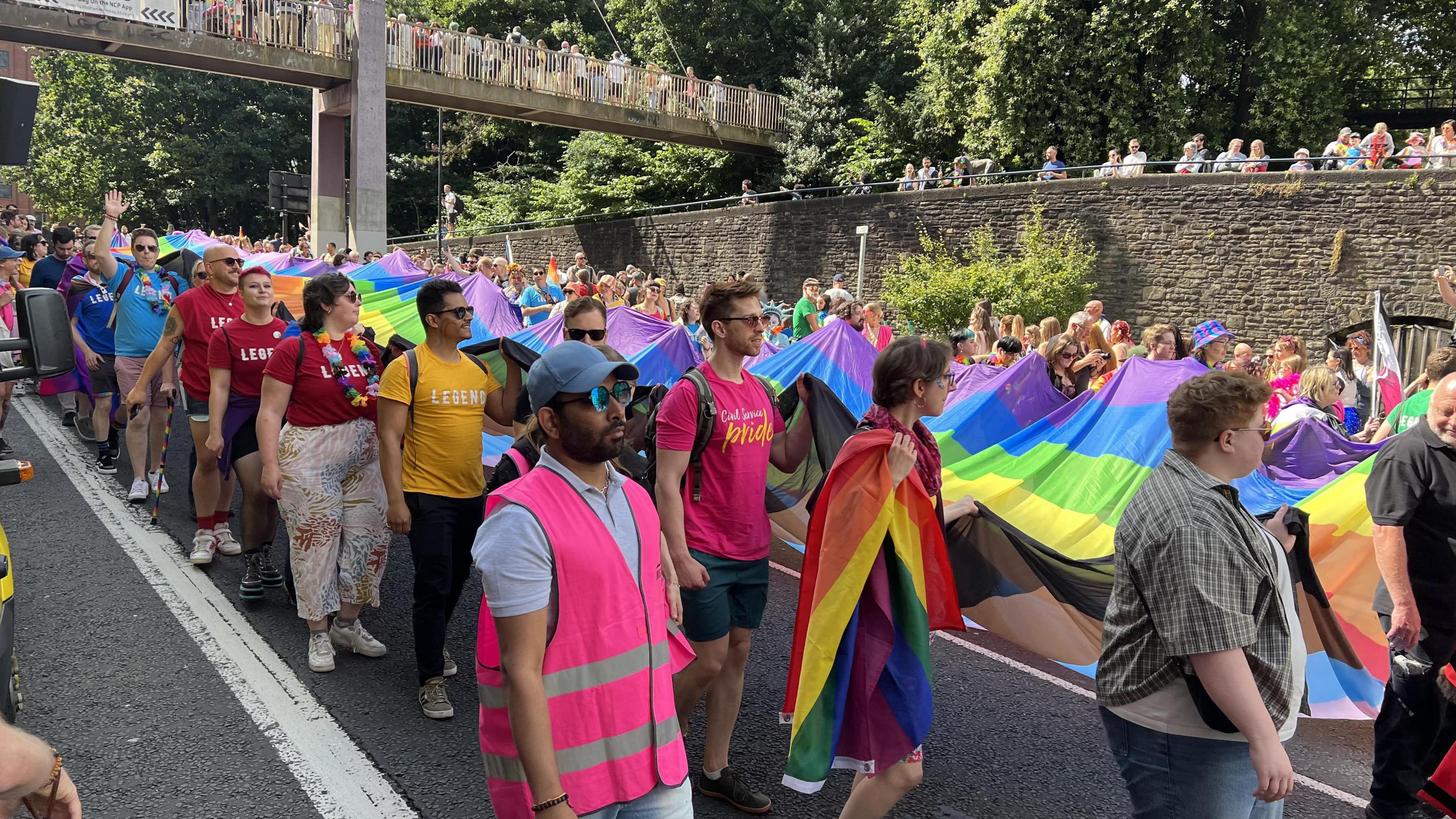 Crowds taking part in the Pride march in 2024. People are walking in the road and are holding a long rainbow coloured flag.