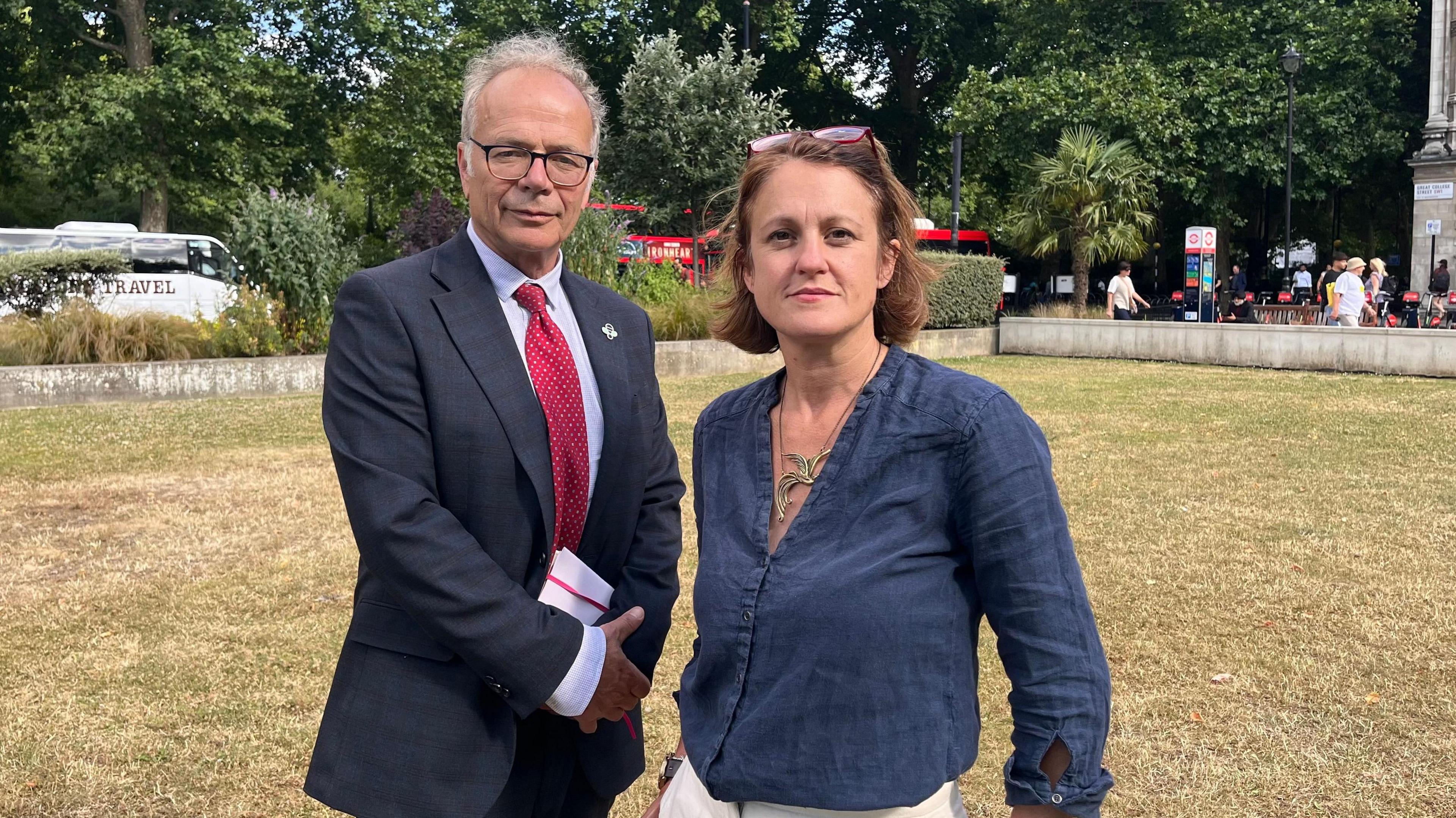 A man and a woman pose for a photo. He wears a suit with a red tie and she wears white trousers and a dark blue shirt. They both look at the camera.
