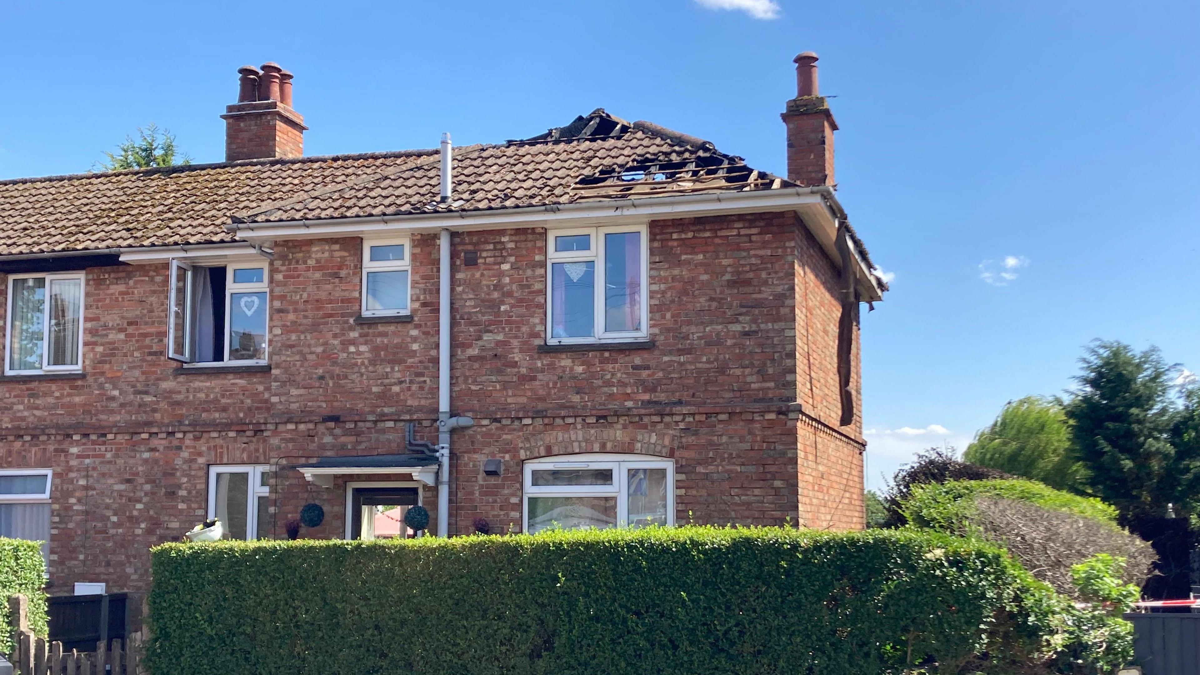 Brick house with a hole in the roof and two chimneys. There are five windows and a door plus two windows on the neighbouring property. There is a privet hedge in front of the house and trees to the side. It is a sunny day.
