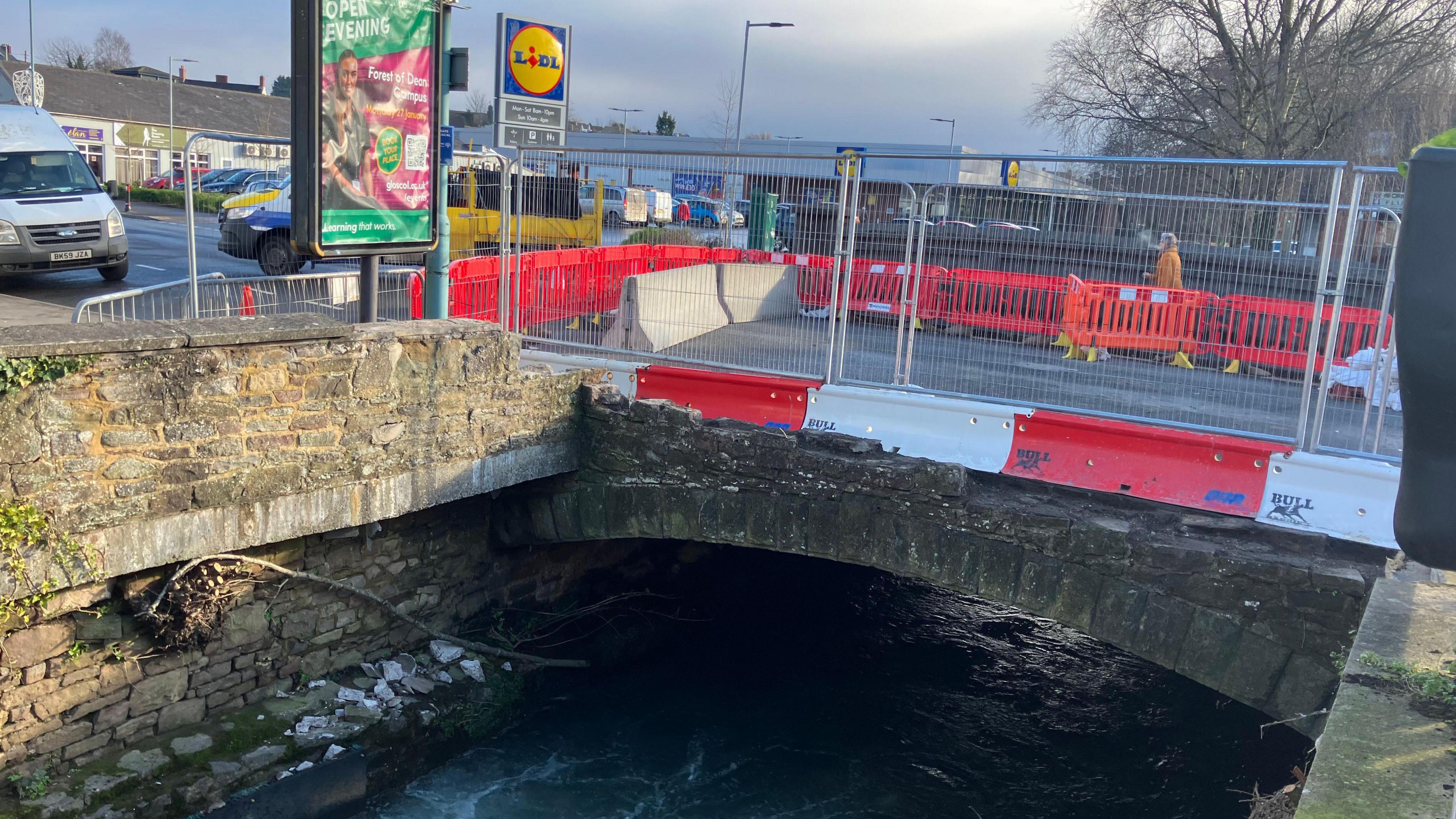 Grey fences and red and white barriers are blocking access to a two-lane road bridge. A section of the side of the bridge is missing, meaning there is a sheer drop down to the water below.