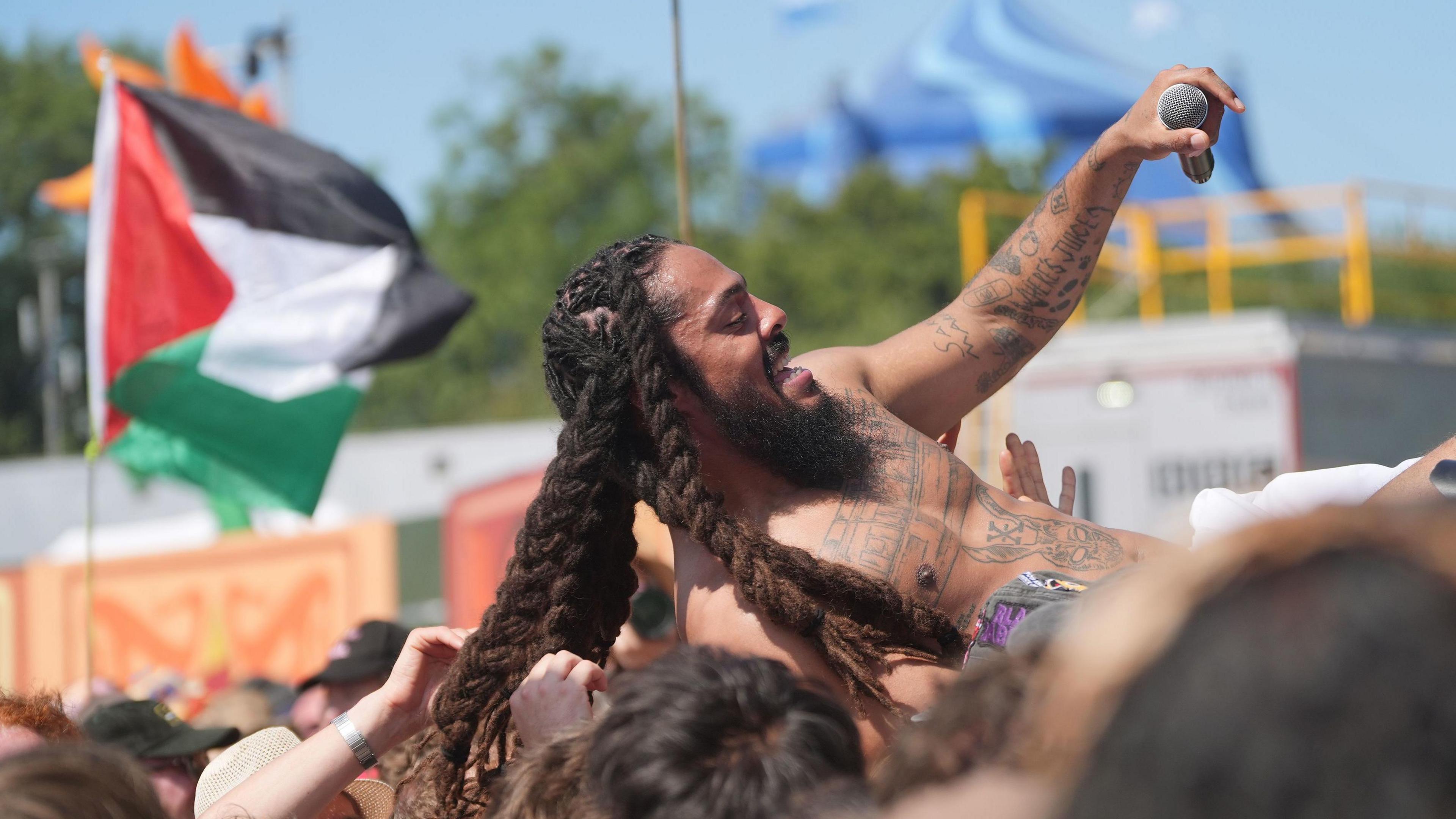 A man with tattoos and dreadlocks crowd surfs during an open air concert. A Palestinian flag flies in the background.