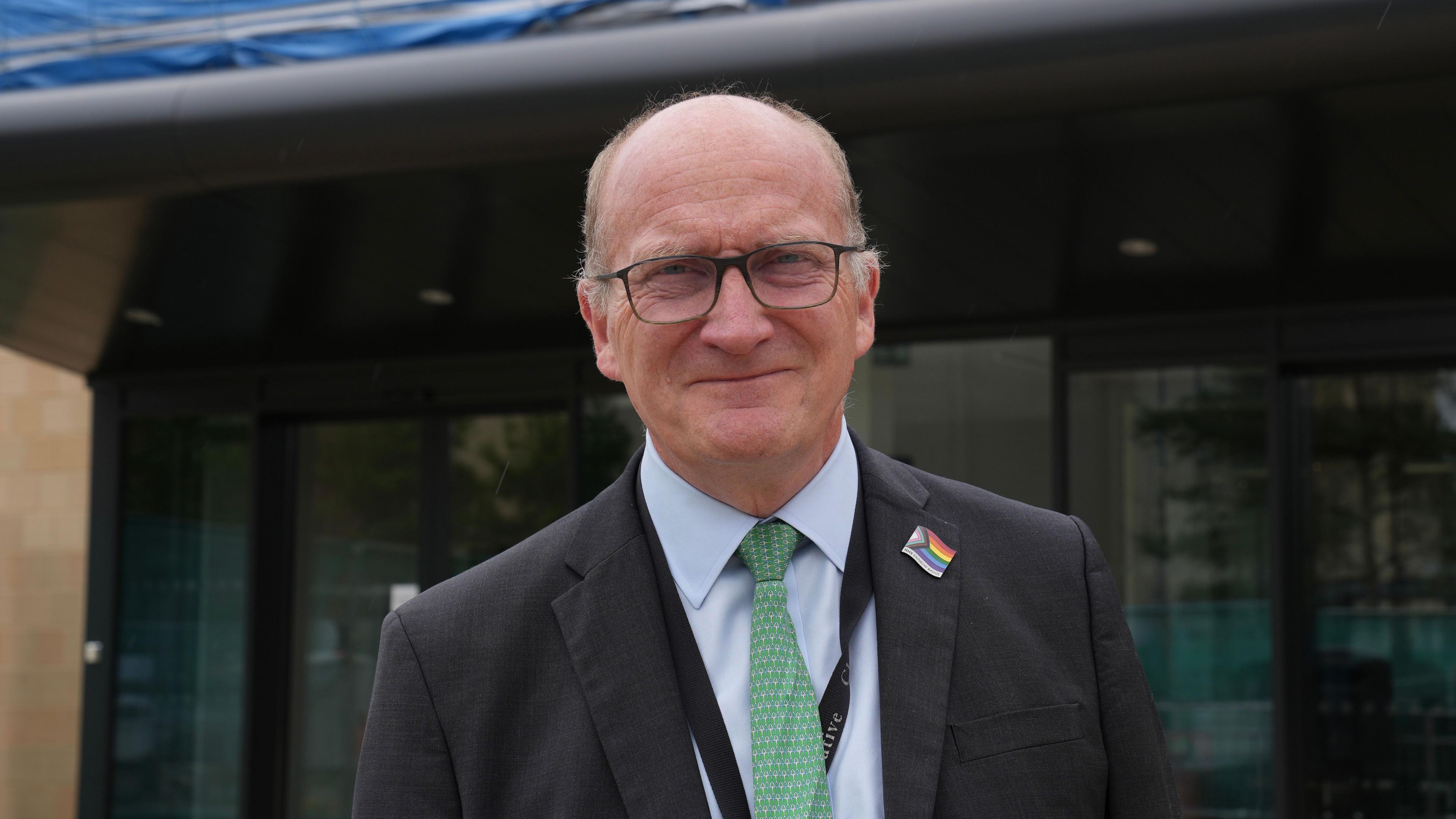 Nick Hulme smiles at the camera standing outside a hospital building. He is largely bald and is wearing glasses. He wears a black suit with a light blue shirt underneath and green tie. He wears a Pride flag pin badge on his chest.