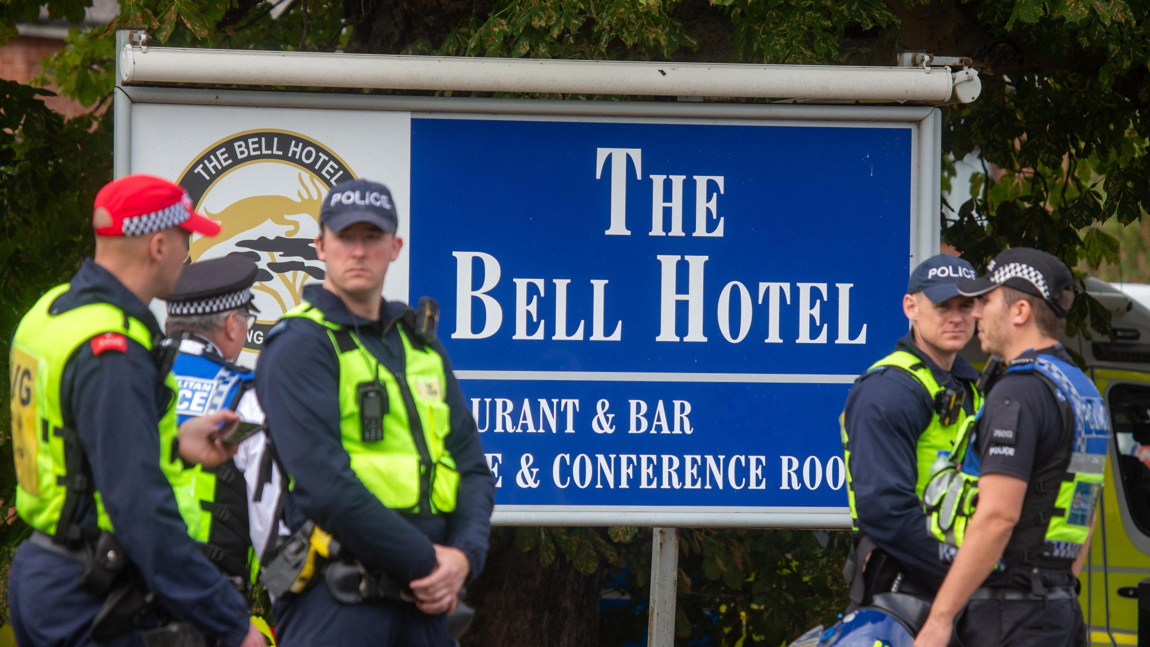 Police officers wearing yellow hi-vis jackets and caps standing in front of a large blue and white sign that says "The Bell Hotel".