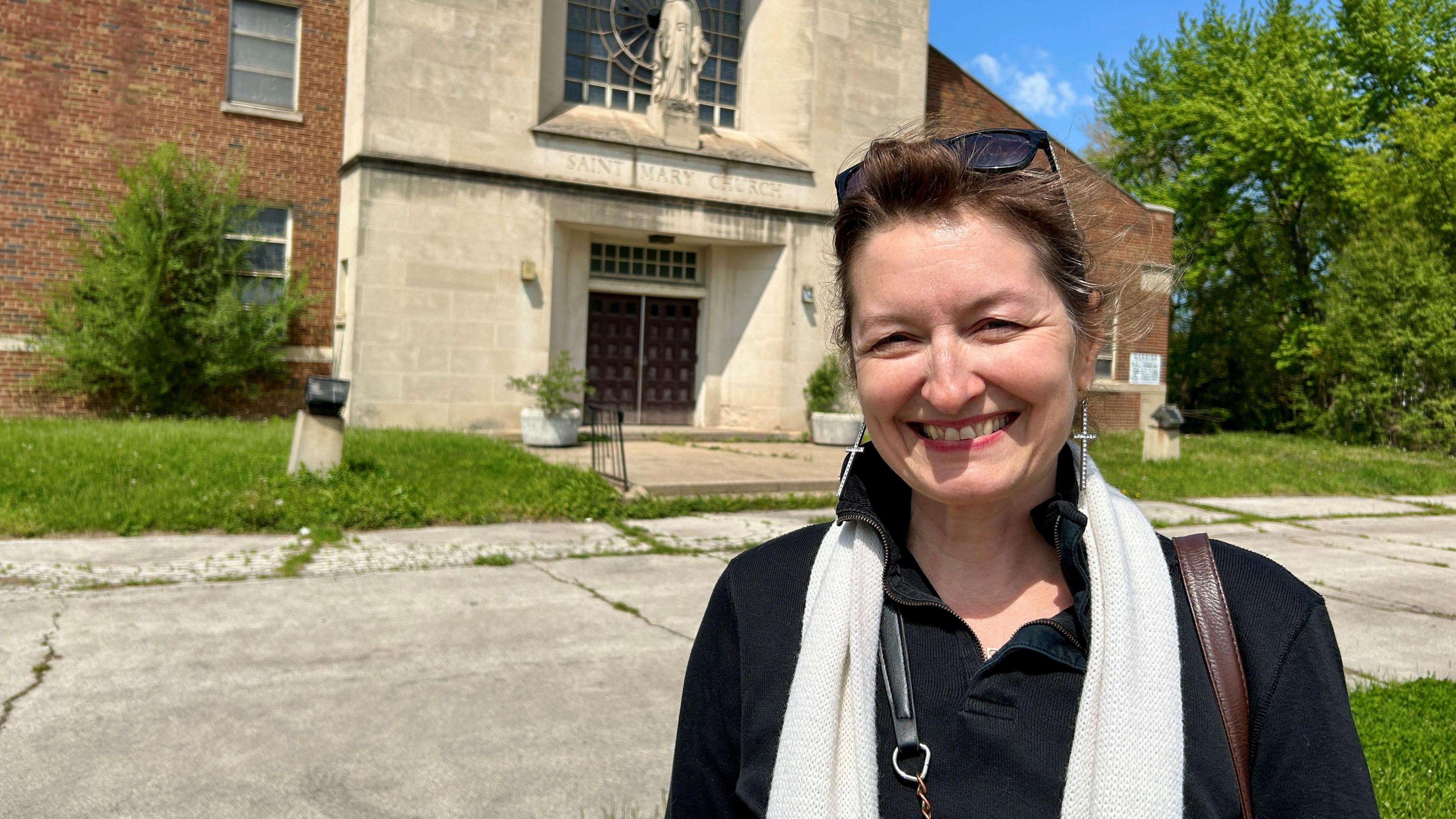 Mary Simons smiling outside of a stone fronted church with weeds in the background.
