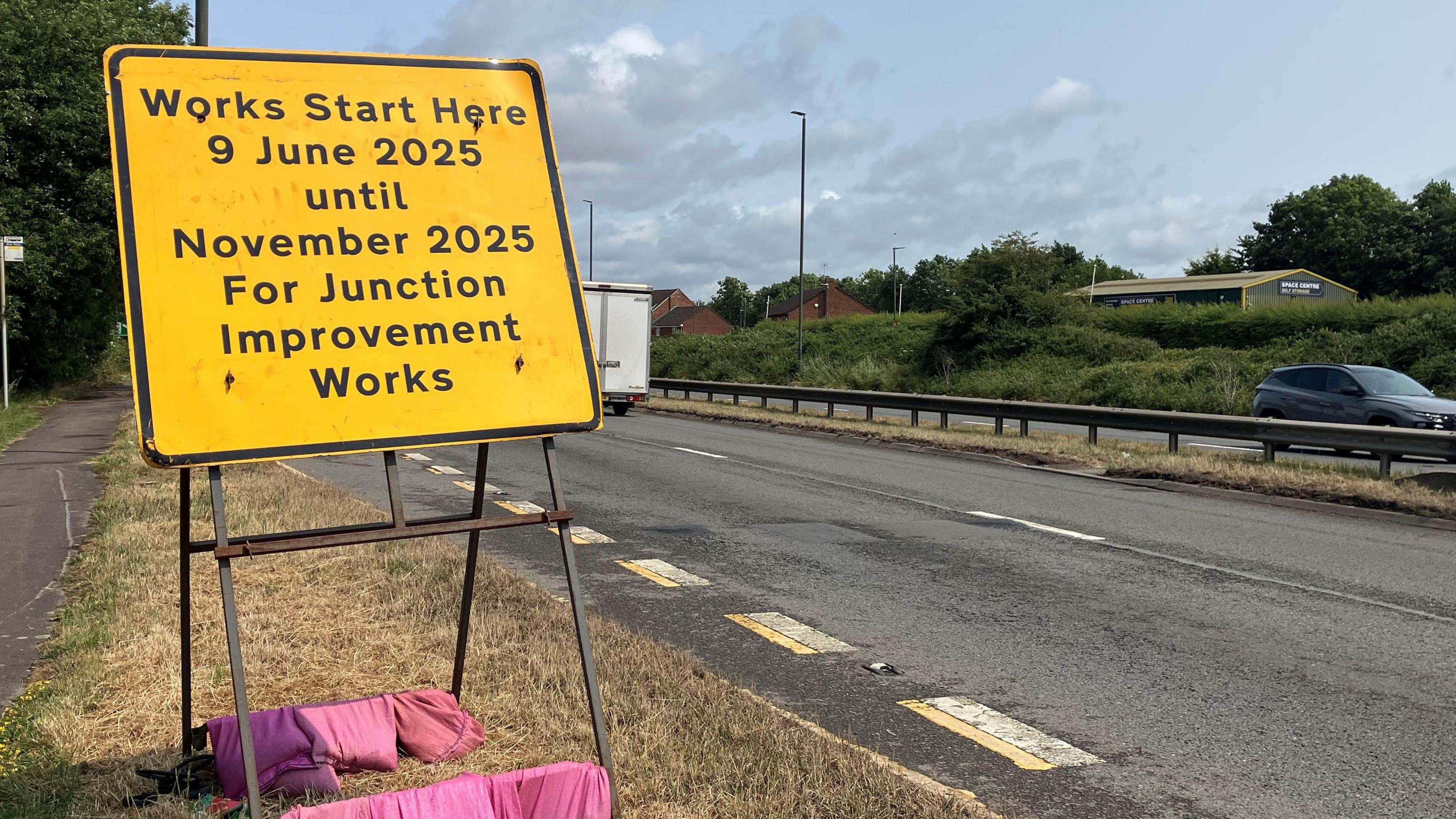 A yellow roadworks sign with 'work starts here 9 June 2025 until November 2025 for junction improvement works'. 
The sign is being held in position by a number of pink sand bags at the base. The sign is on the side of a dual carriageway. 