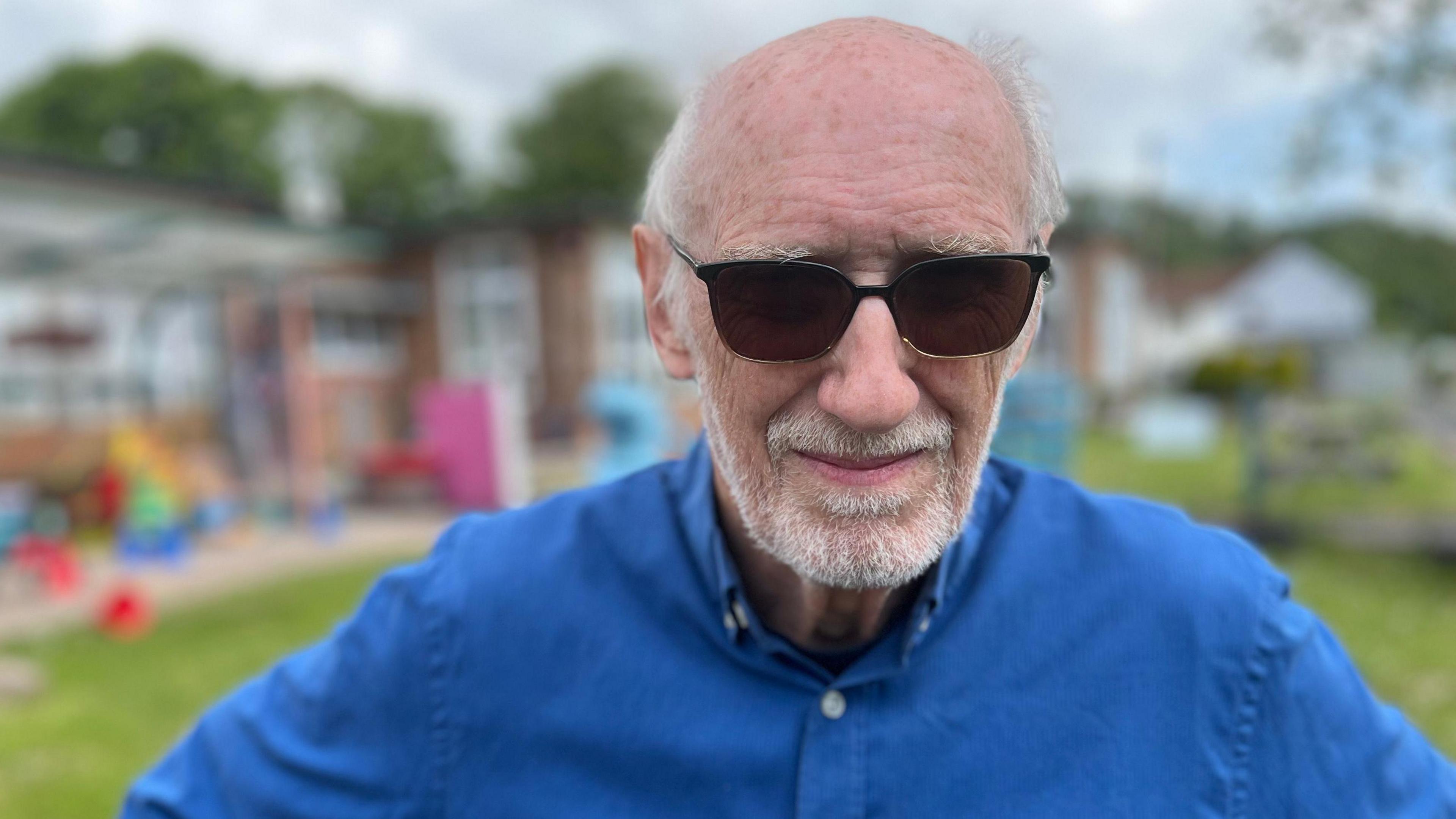 A man looking at the camera. He is wearing a blue shirt and has a grey beard and moustache, it is stubbly. He has dark shades on and behind him is the school, which is slightly out of focus.