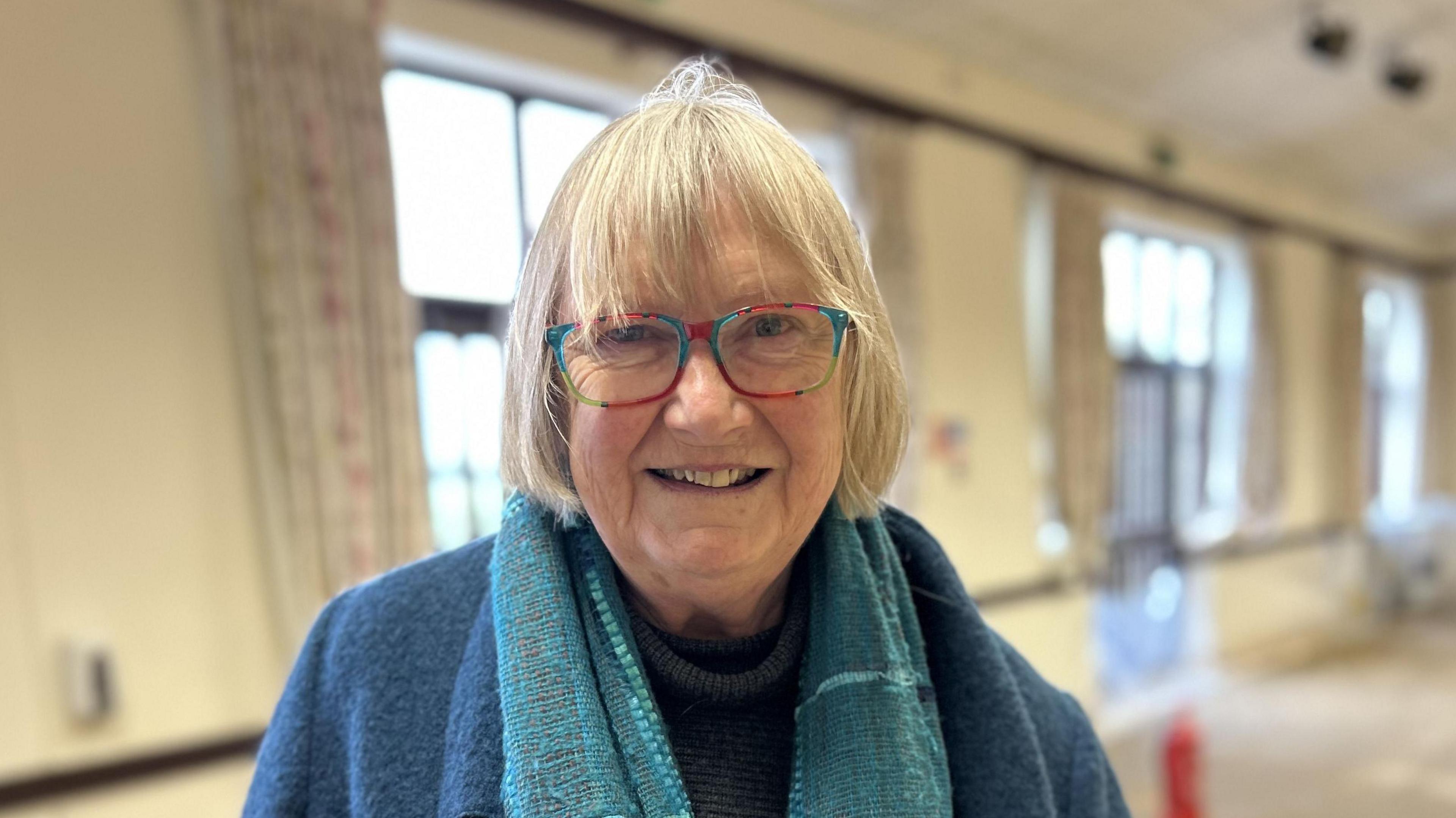 Sally Wilks with grey hair stands in the village hall. She has red and blue-framed glasses, and is wearing a turquoise scarf and navy blue jacket.