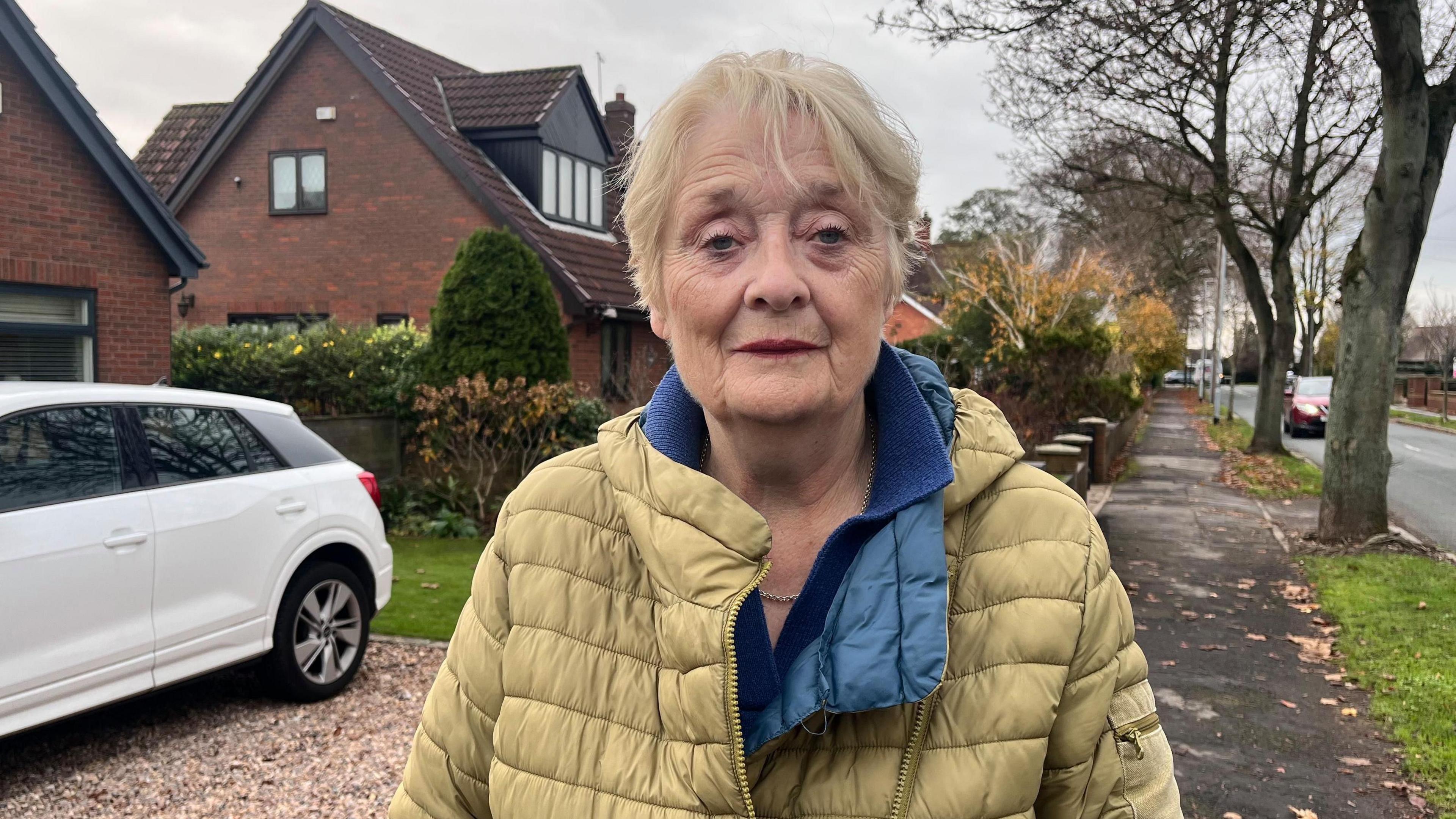 Mary stands outside her home in Cottingham wearing a yellow jacket and blue top and stares directly at the camera, looking concerned. Behind her is a gravel drive and white car.