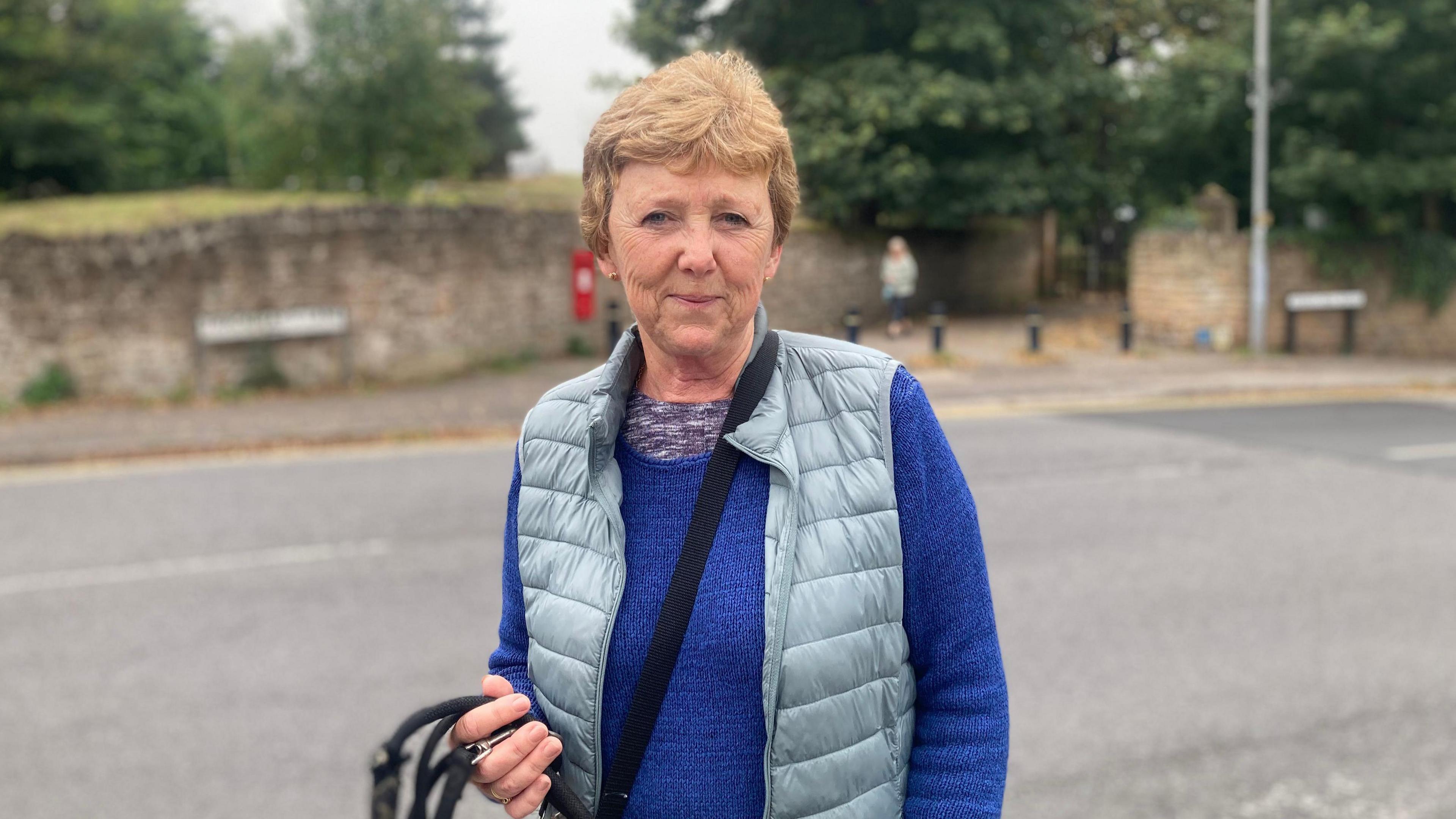 A woman wearing a blue jumper and brown gilet stands in front of a road. On the other side of the road is an old stone wall and a postbox.