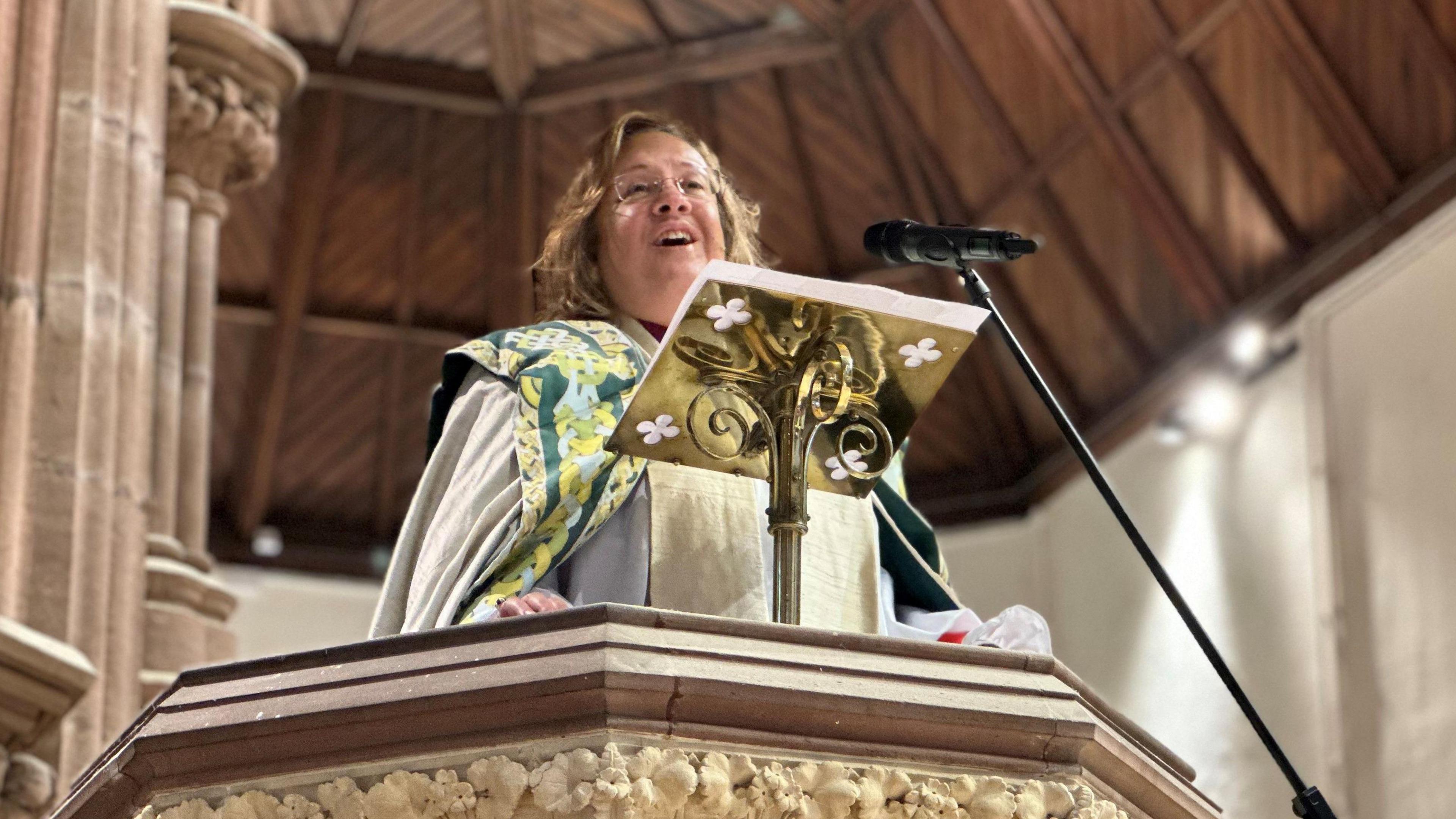 The Right Reverend Patricia Hillas in the pulpit in Cathedral Isle of Man. She is wearing a vestment of beige, green and yellow, and there is a gold and a black microphone in front of her