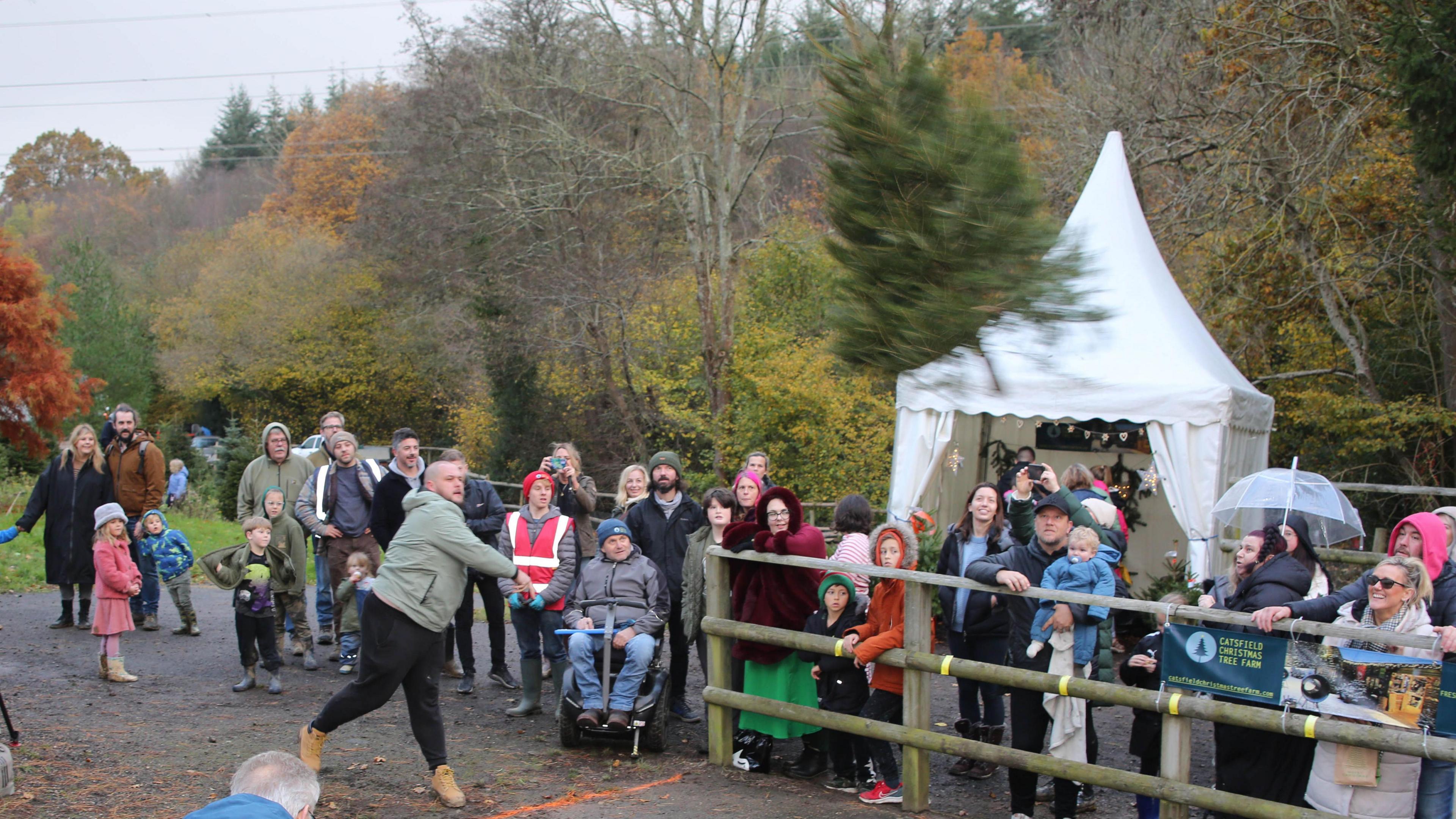 Roughly two dozen people stood behind a wooden fence next to a road. They watch as a man throws a Christmas tree up the road. Behind them is a white gazebo and woodland.