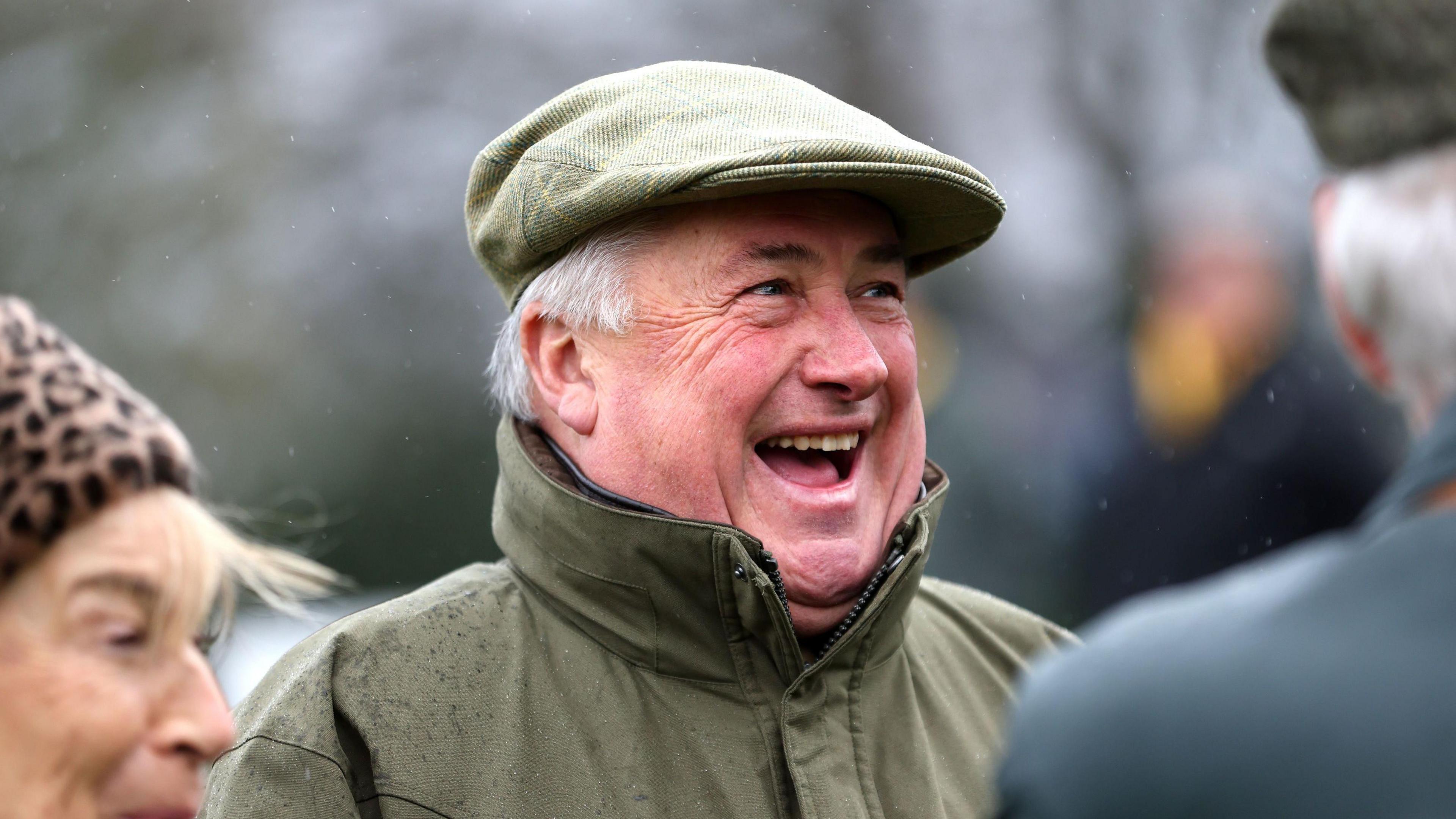 Paul Nicholls smiling with his mouth open, wearing a khaki coloured coat and cap