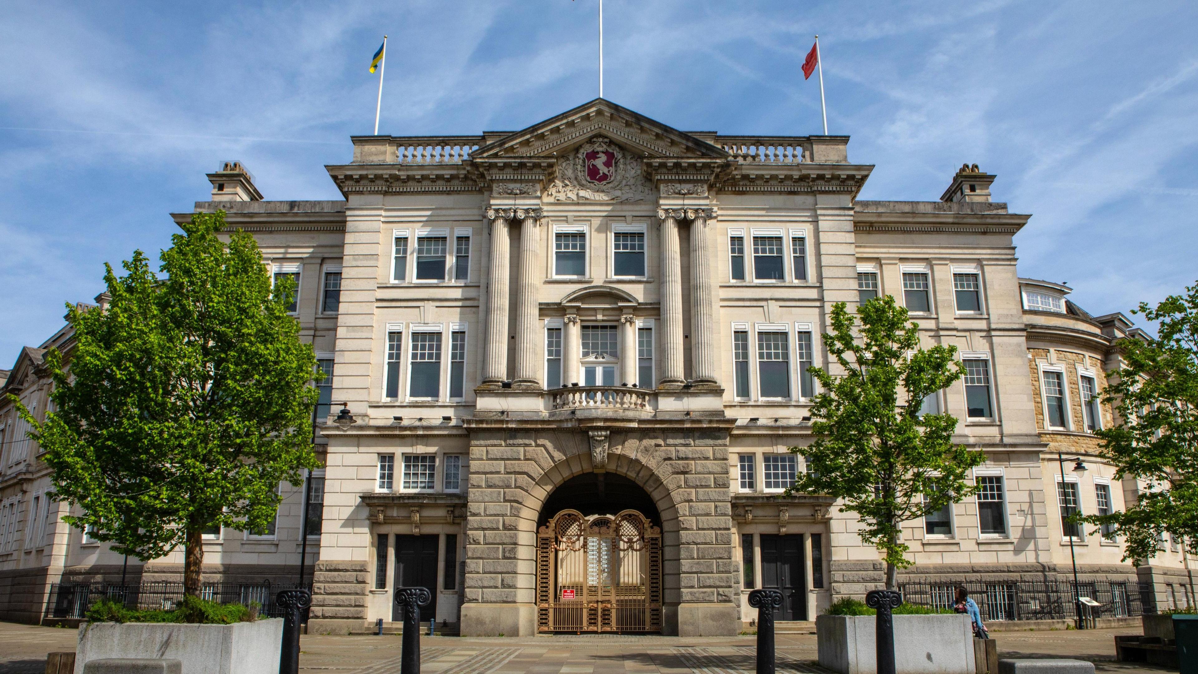 The outer facade of County Hall in Maidstone, a late Victorian building in the neo-Palladian style in white Portland stone, with an arch of bricks supporting two pairs of pillars, and high windows. Three trees in planters and two traffic bollards flank the entrance, with three flagpoles, two of them showing flags, one the flag of Kent, the other the flag of Ukraine.