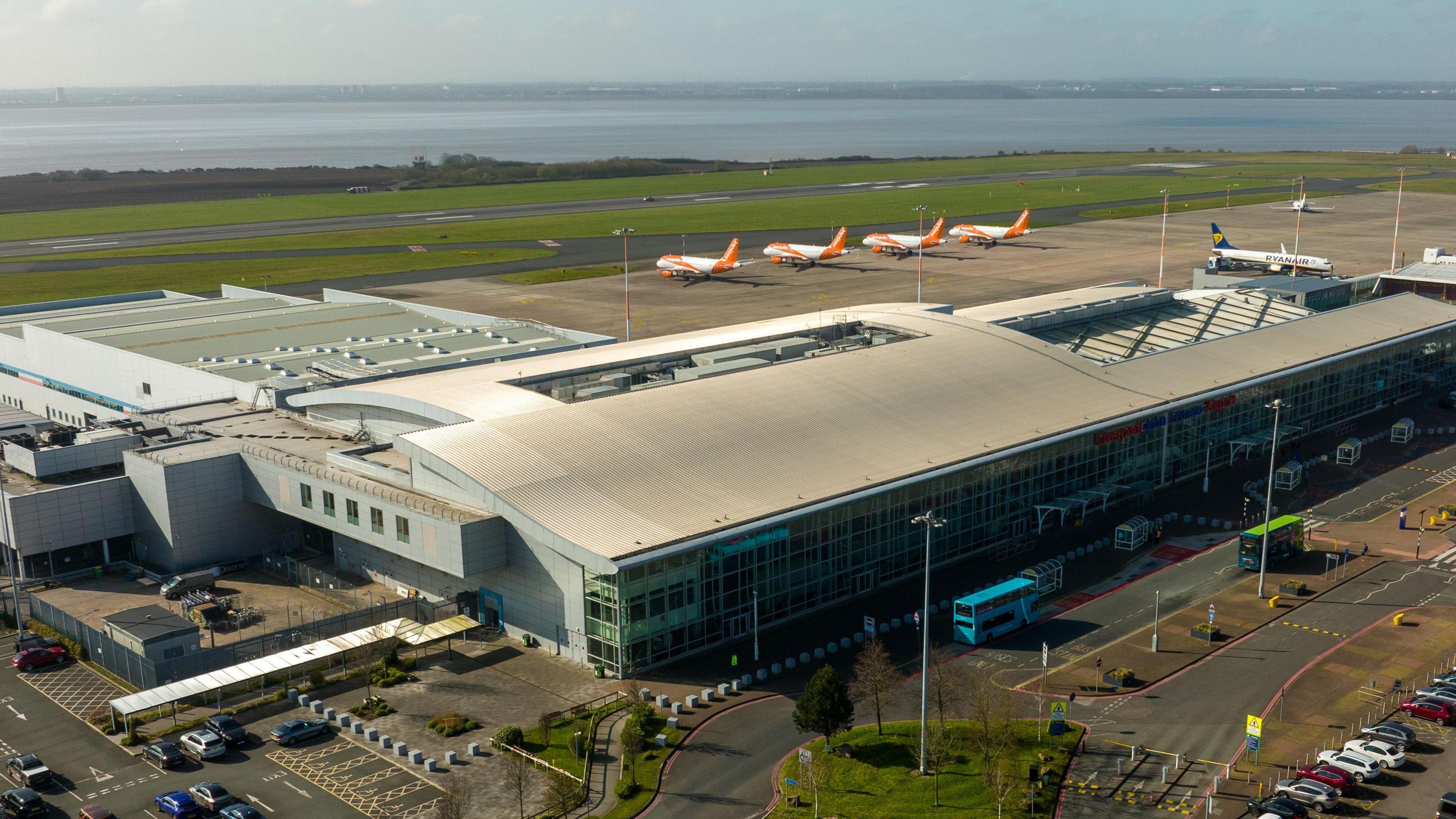 An aerial view of an airport terminal nuilding, with orange and white passenger aircraft visible parked next to a runway.