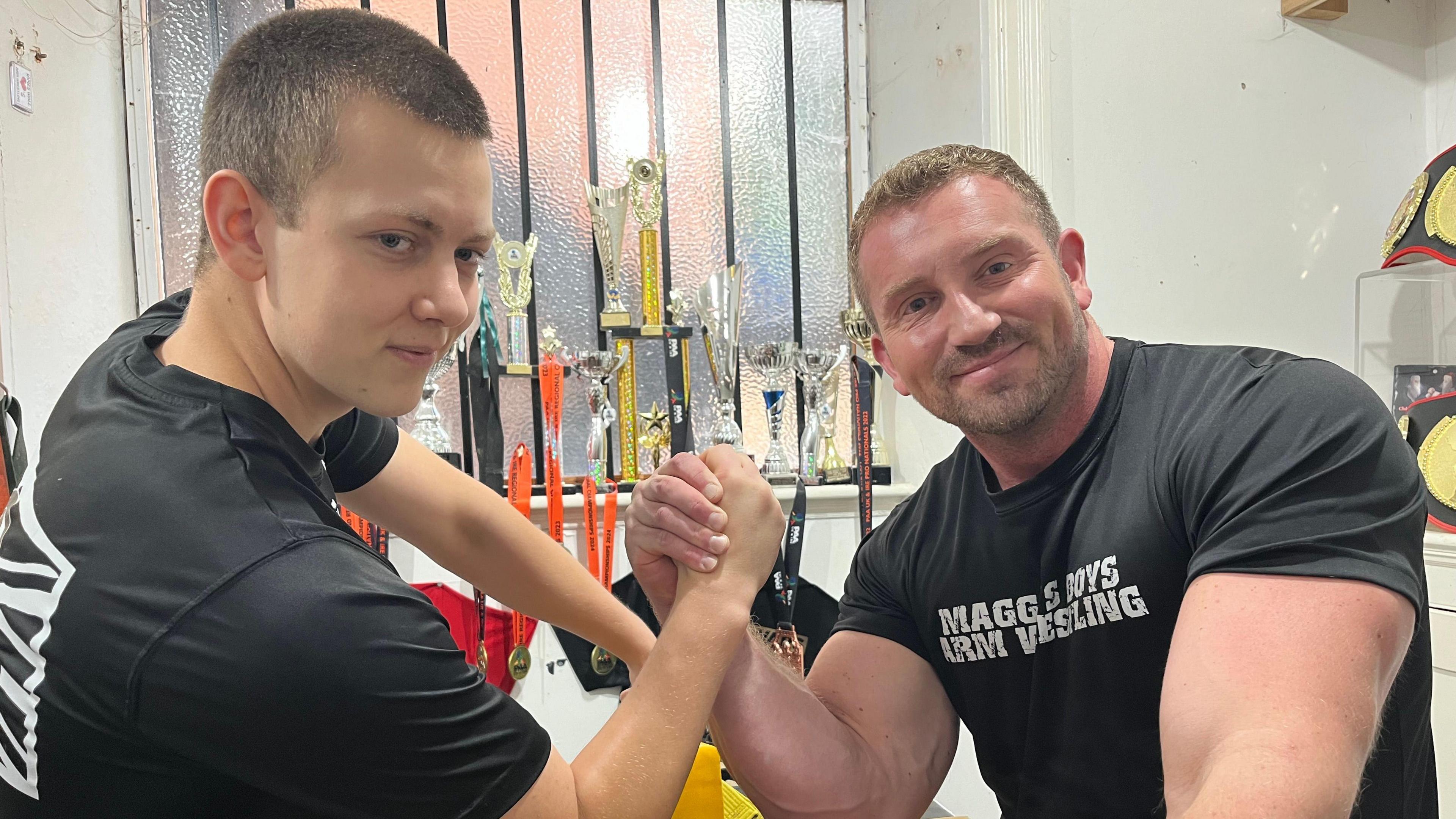 Jack Orme and James "Brick" Wall with hands links as the arm-wrestle and look into the camera. Both are wearing black T-shirts. 