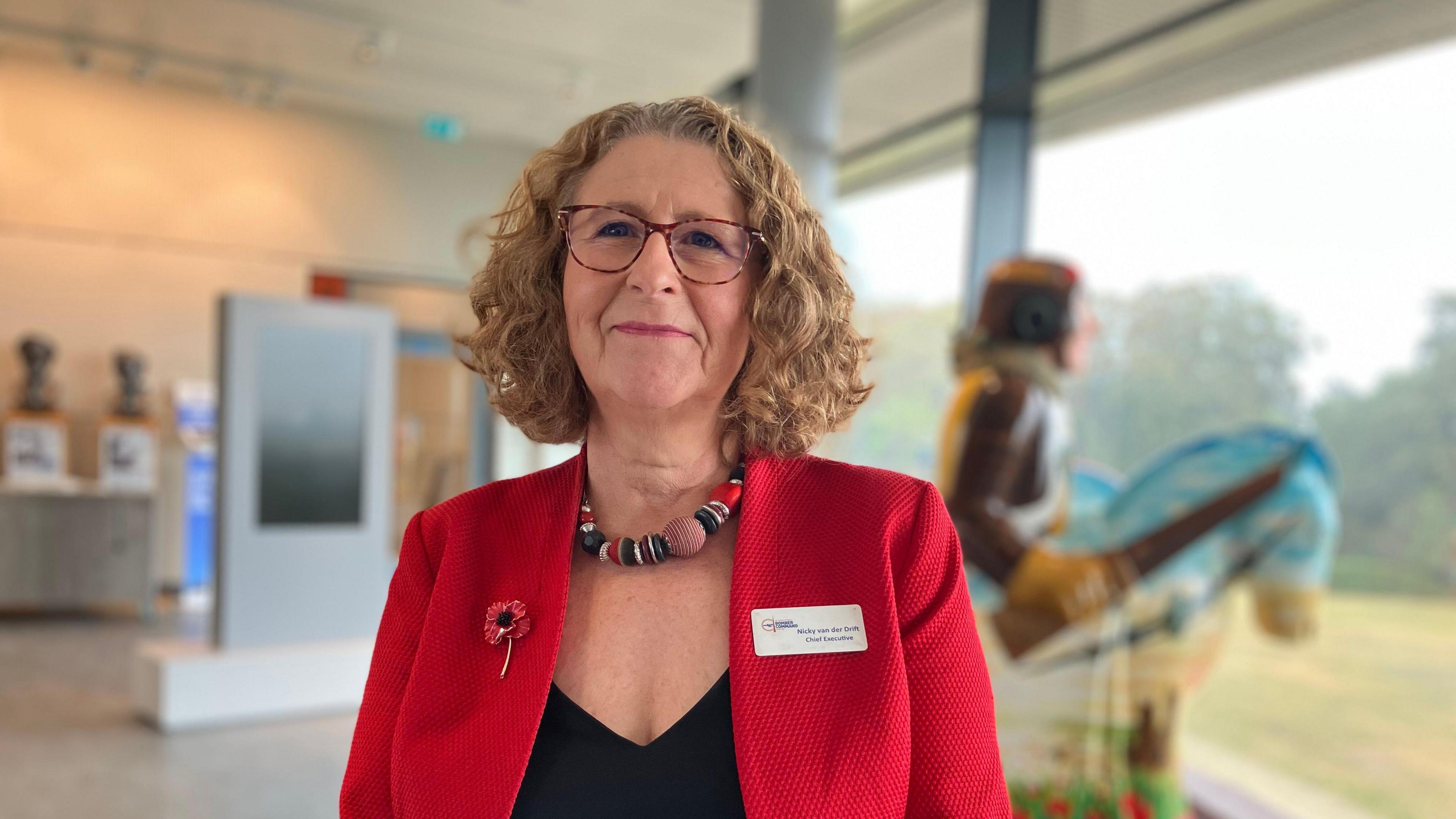 Nicky van der Drift with brown shoulder length hair, glasses, red jacket, black top, a poppy badge and a name badge. She is smiling. She has a beaded necklace. There is a blurred background behind her that looks like a visitor centre. 