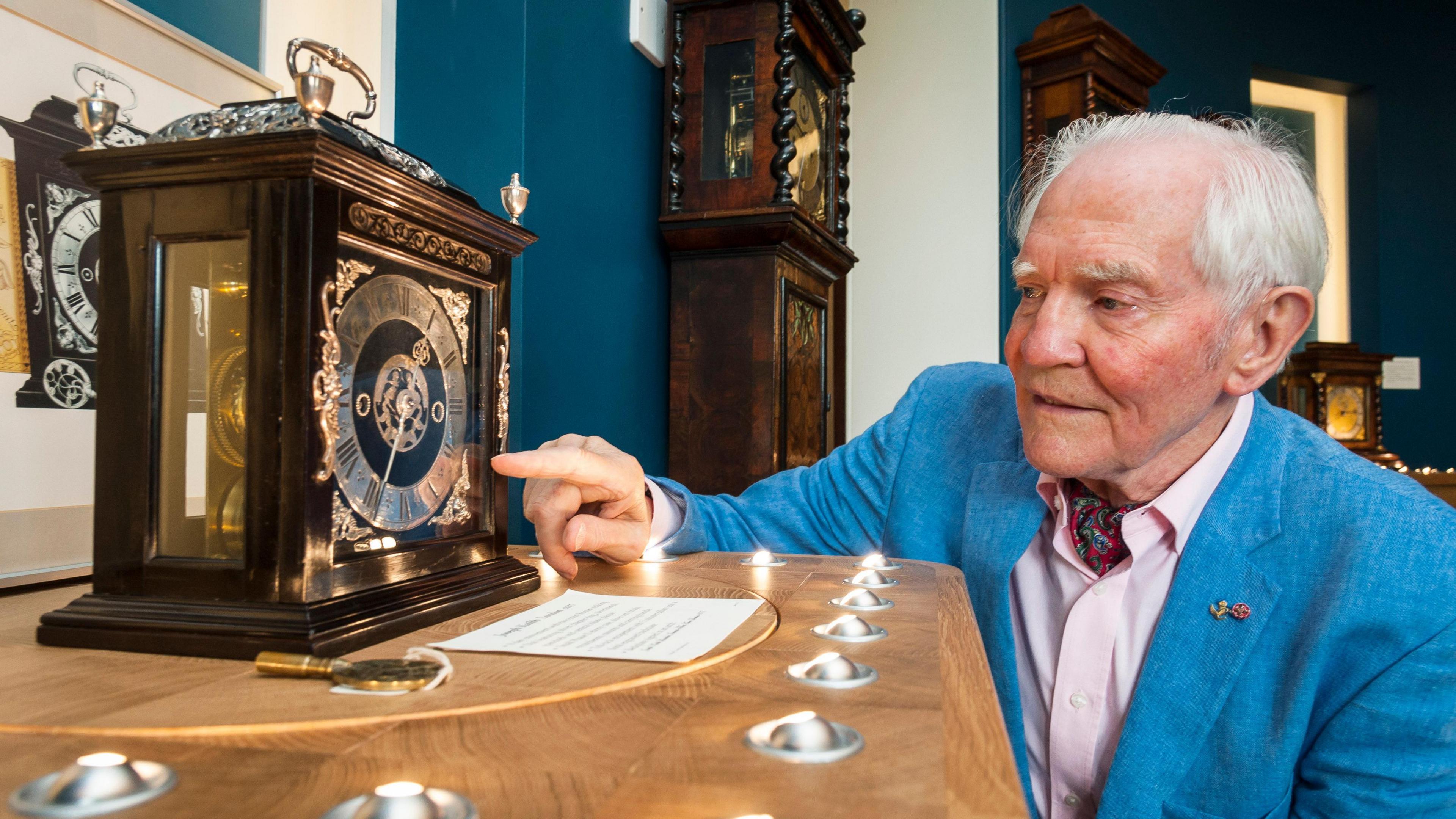 Dr Taylor with one of his historic clocks. He is wearing a blue jacket a pointing at the decorative piece with is on a wooden table.