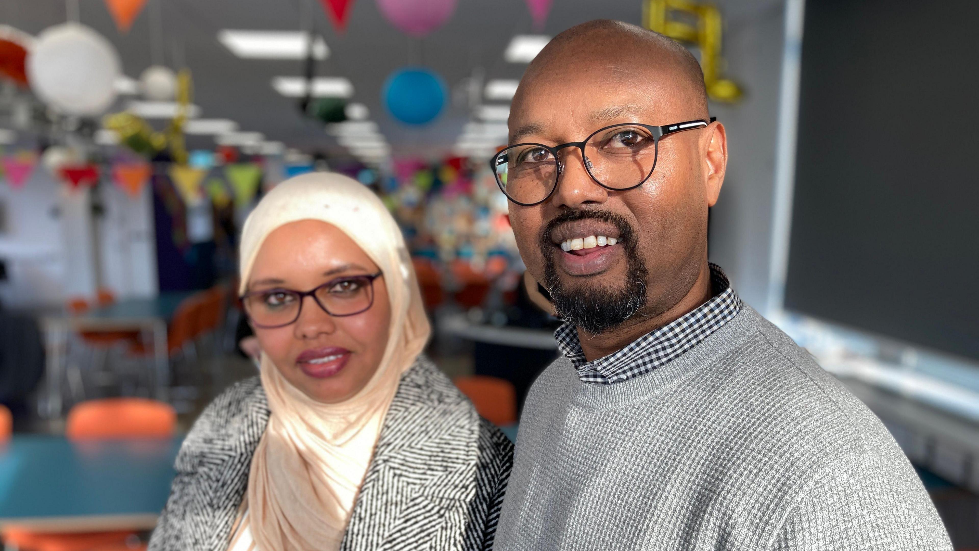 Abdullahi Abdi wearing a blue and white checked collared shirt underneath a waffle patterned grey jumper. He is wearing black framed glasses, has a shaved head and a dark goatee style beard, and is smiling at the camera. Beside him is a woman wearing a black and white patterned coat, a cream hijab and black framed glasses.