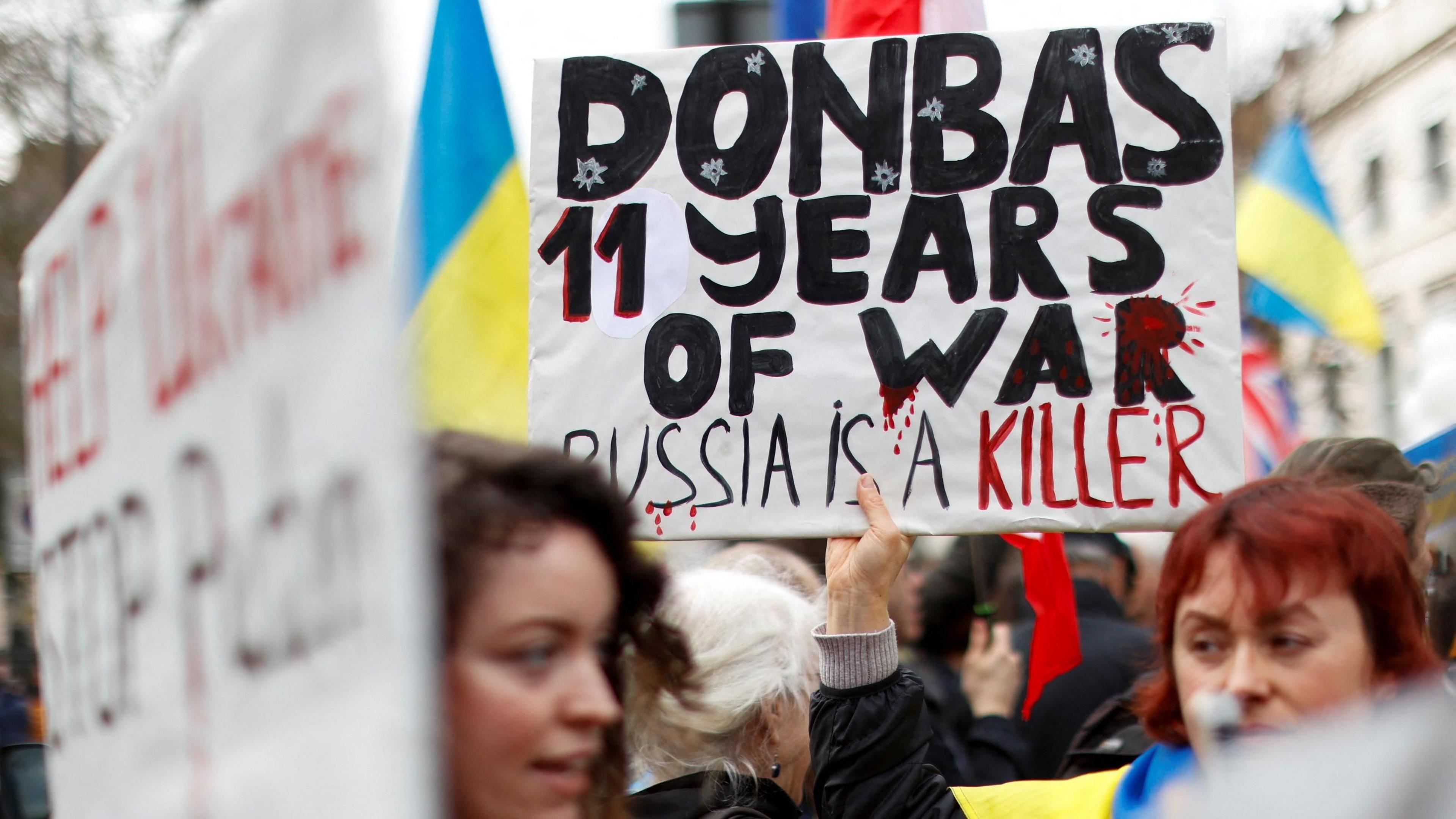 Two women stand in a crowd of protesters, one holding up a poster saying "Donbas 11 years of war, Russia is a killer"