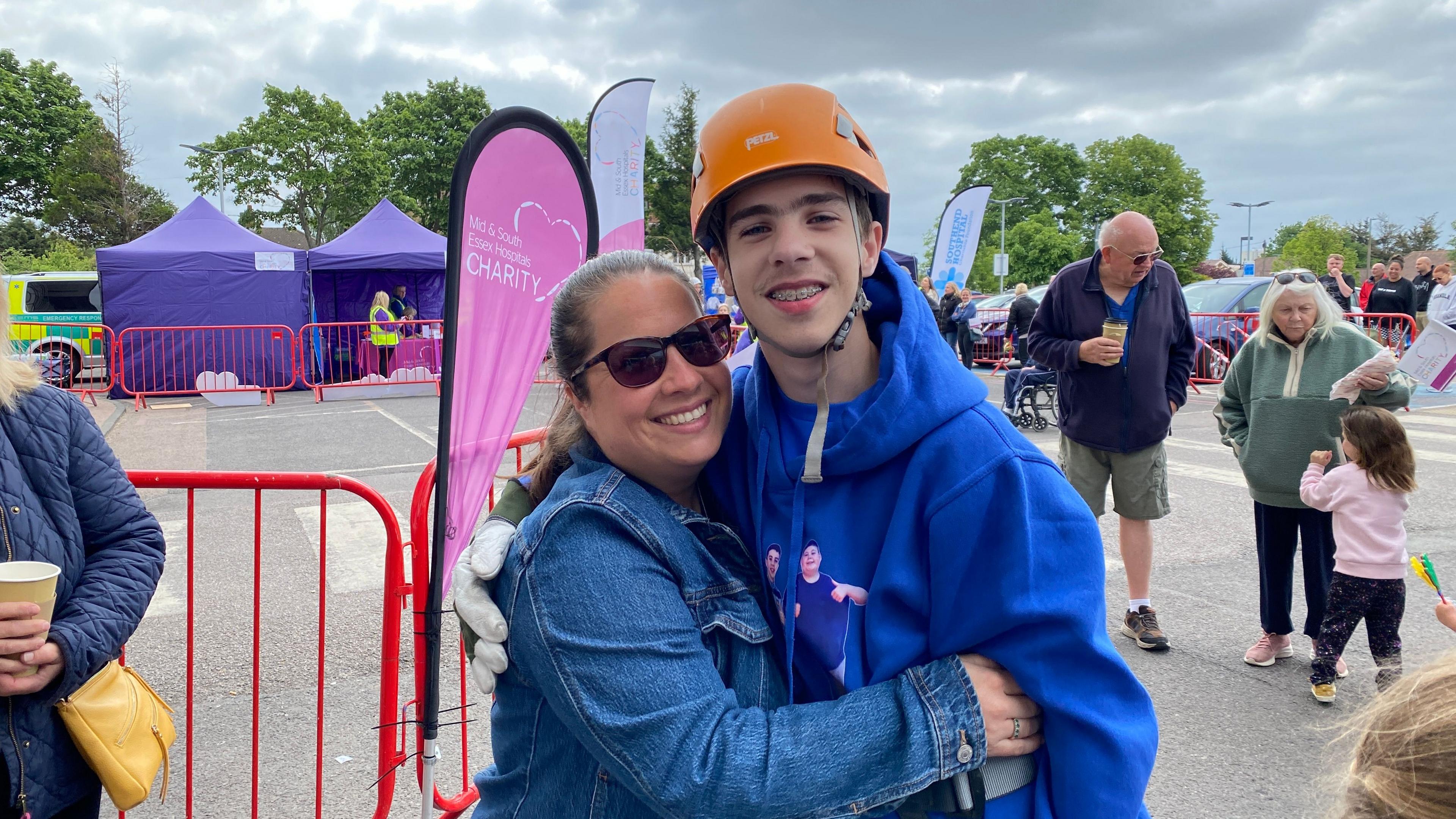 Finley's mum Rhys gives him a hug after he finished the abseil. She wears a blue denim jacket and sunglasses and has her hair tied back. Finley wears his orange helmet and blue hoodie, and smiles at the camera