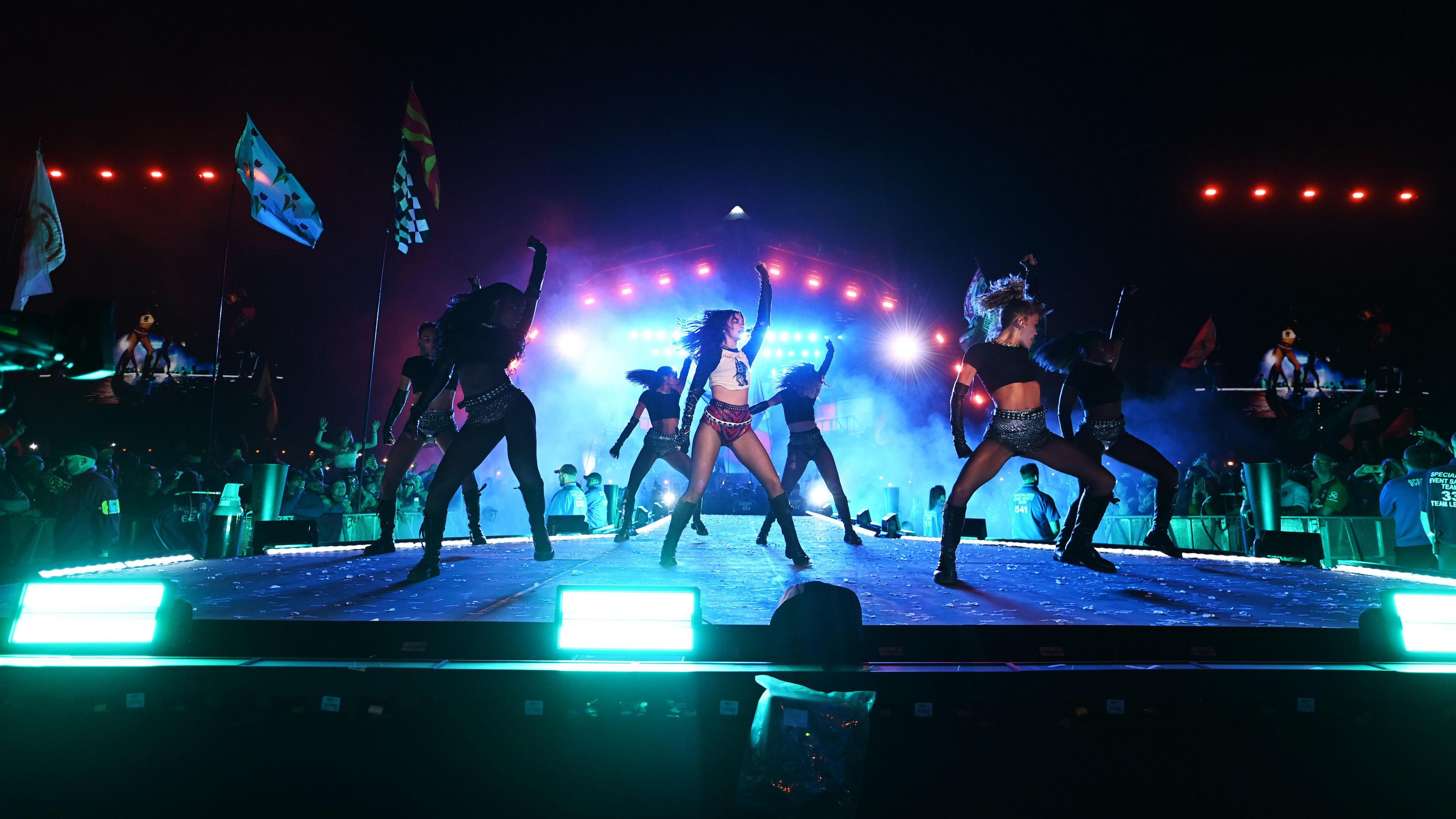 Dua Lipa performing on the Pyramid Stage at Glastonbury Festival on 28 June 2024. She is dancing and has one arm in the air, surrounded by six other dancers.