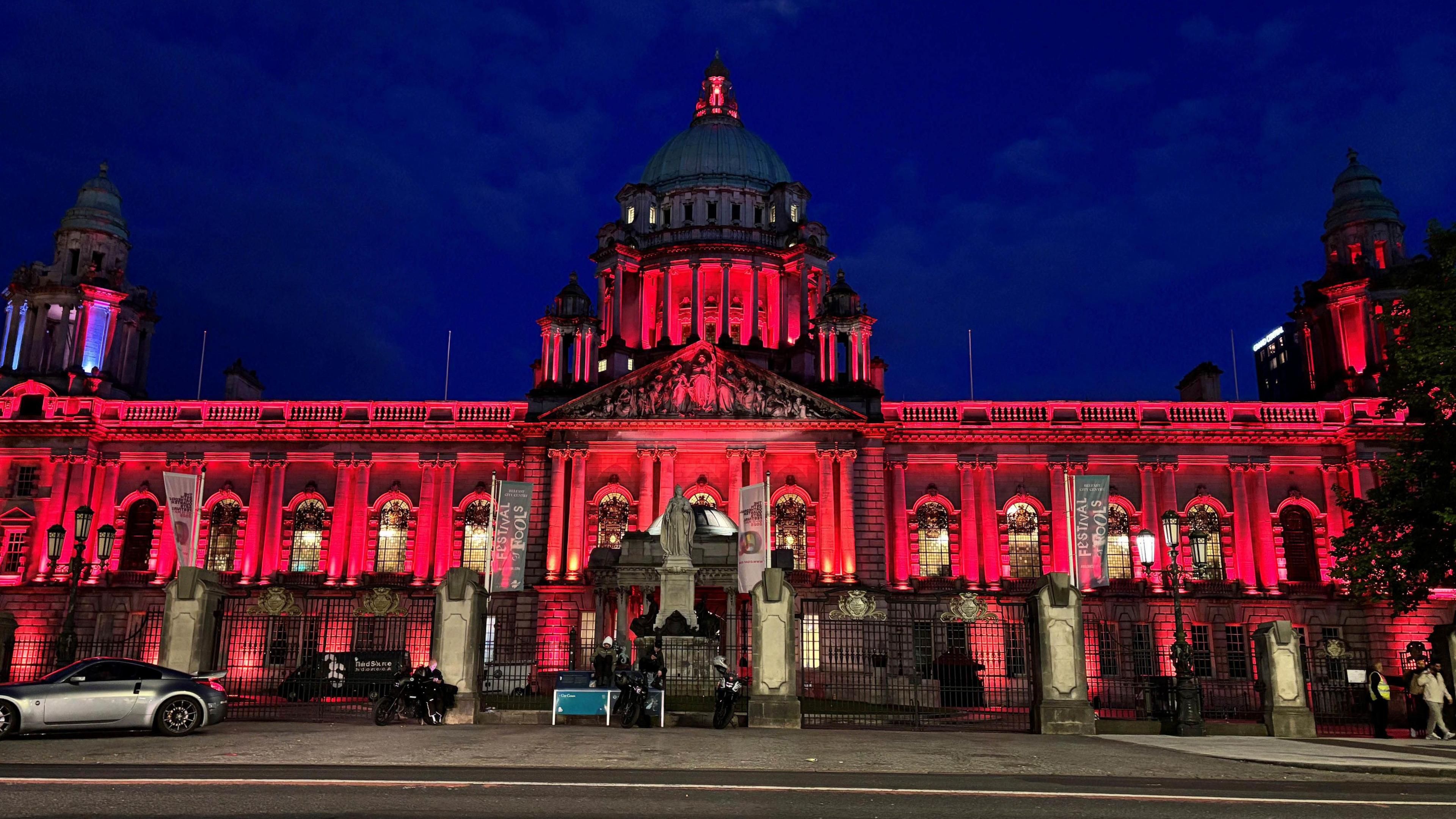 The front of a stone building with domes and lots of windows, lit up red against a night sky