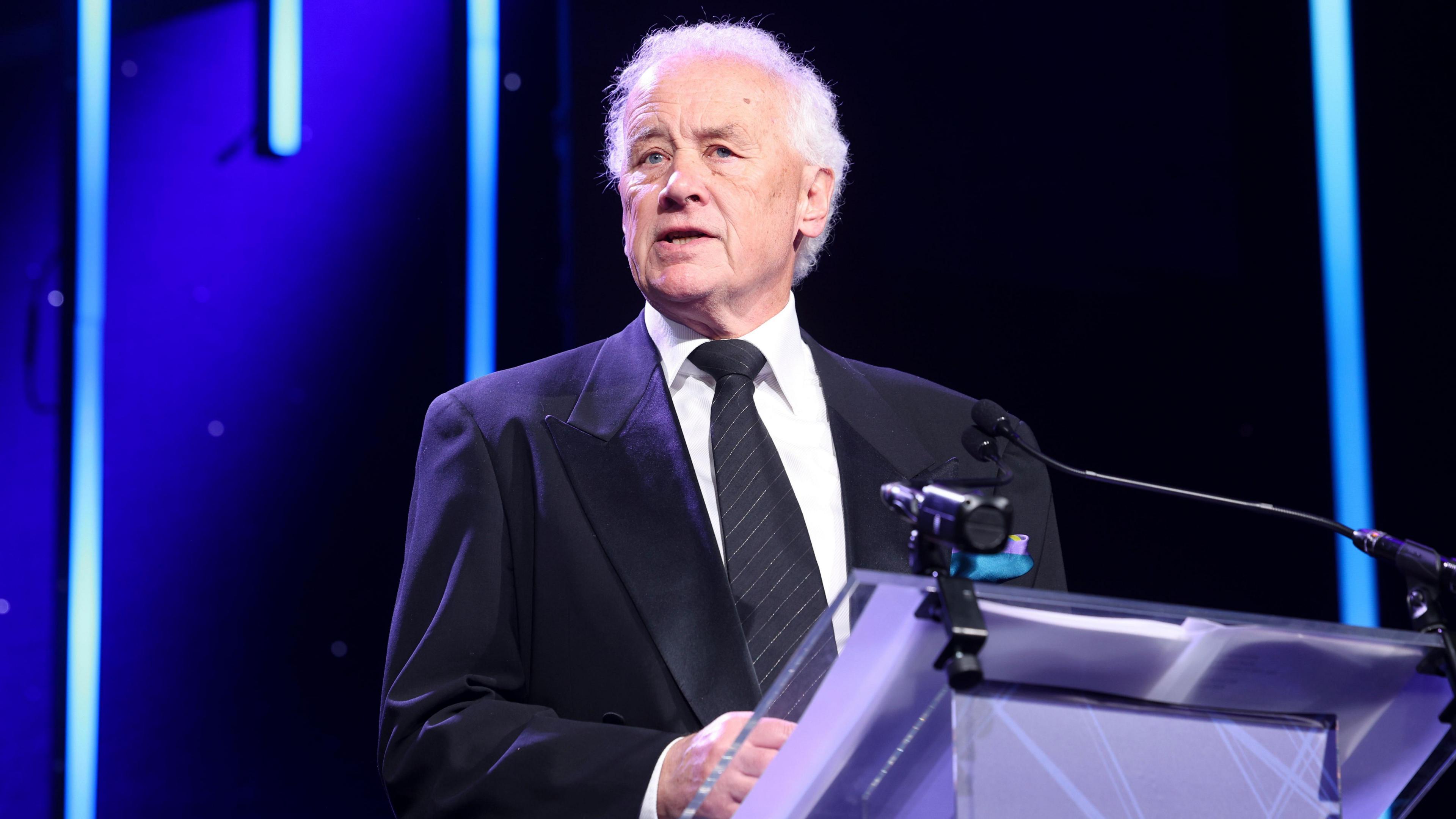 Ricky Parry wearing a blue suit jacket, white shirt and black tie with silver diagonal stripes standing behind a lectern addressing the EFL awards ceremony