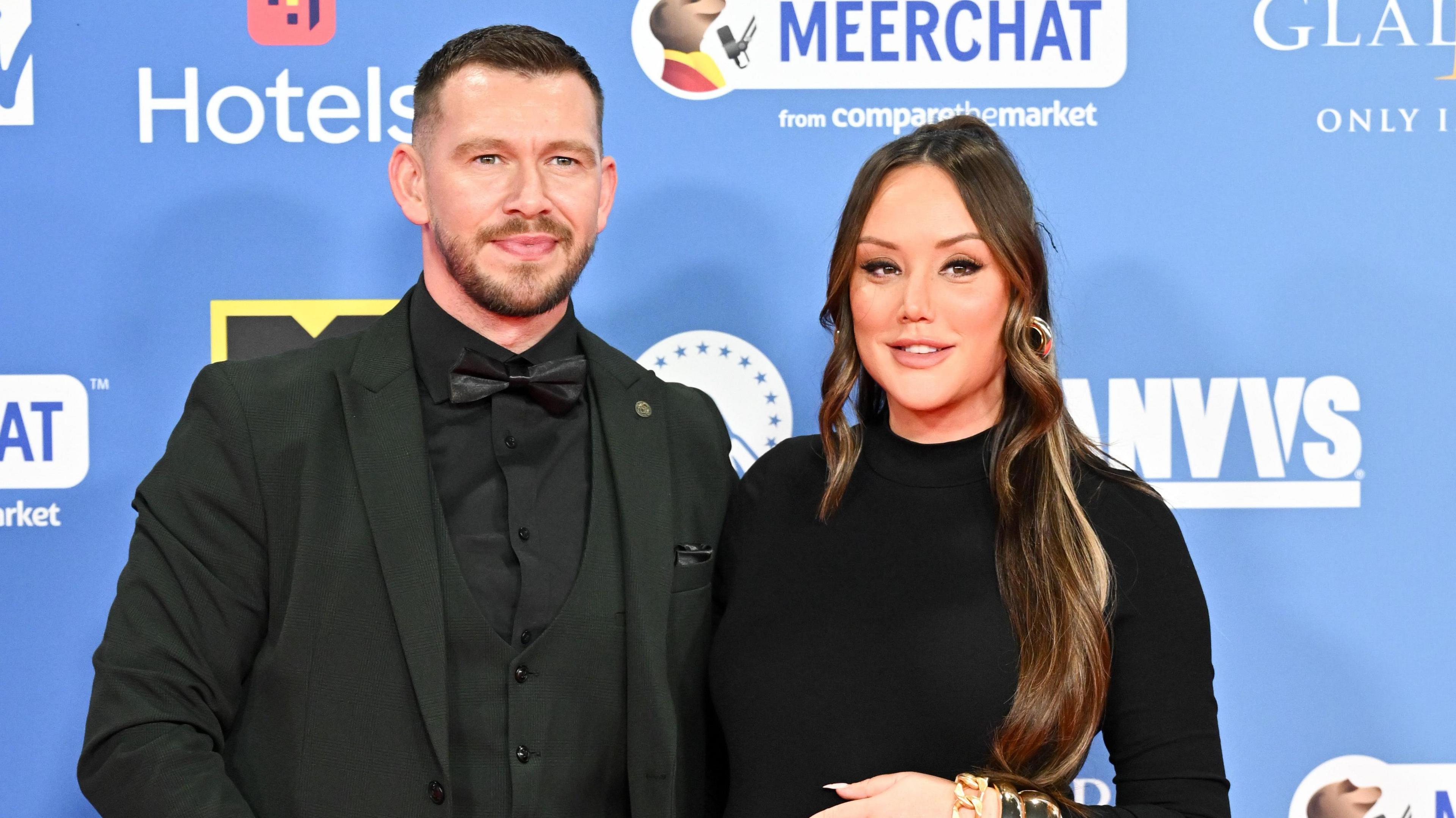 Jake Ankers and Charlotte Crosby smiling on a red carpet. Jake has brown, short hair and is wearing an all-black shirt, suit and bow tie. Charlotte has long, curled brown hair and is wearing a long-sleeved black dress.