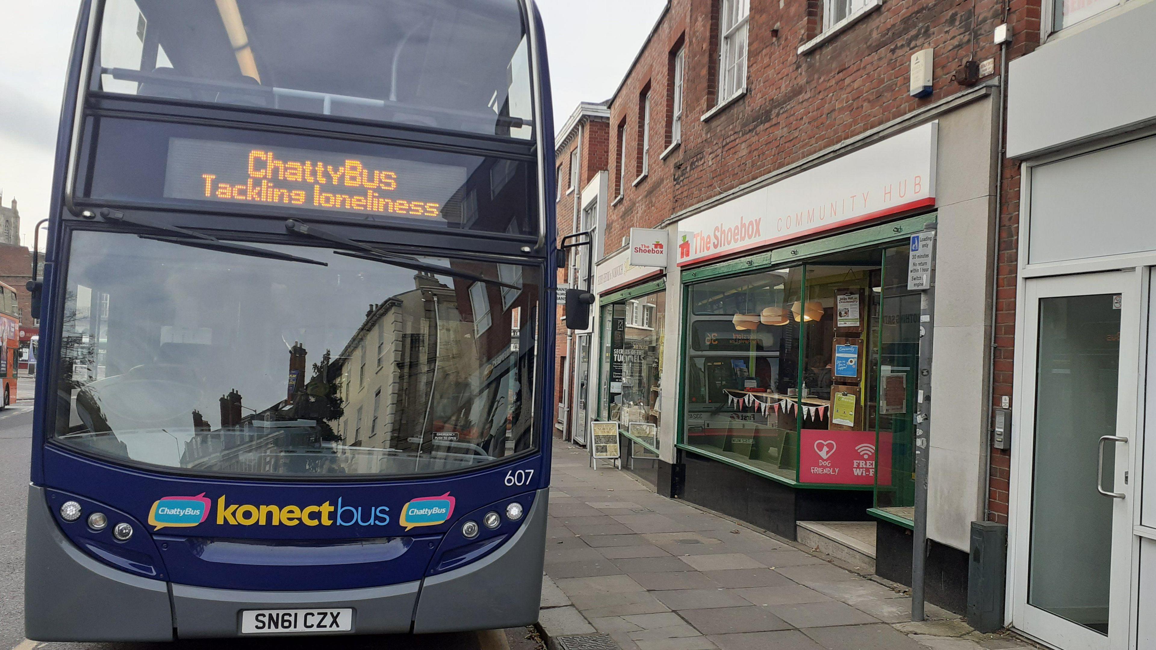 A blue and grey Konectbus. It is parked outside a shop and on the front of the bus it says "Chatty Bus tackling loneliness".