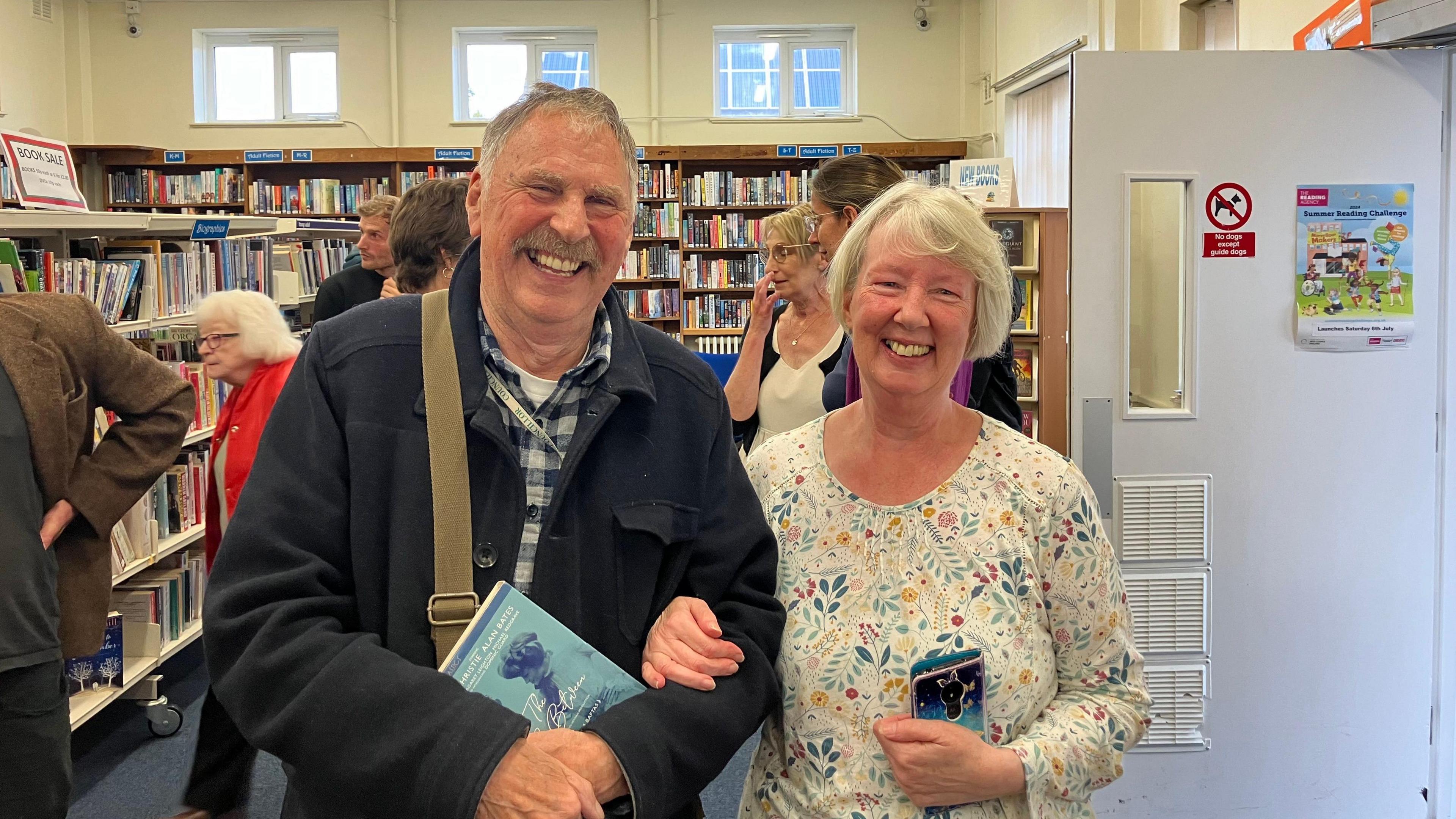 Alan Dowson wearing a navy coat with Phillipa Philips in a white T-shirt with colourful flowers. Both are smiling at the camera and linking arms, while being photographed in the library. 