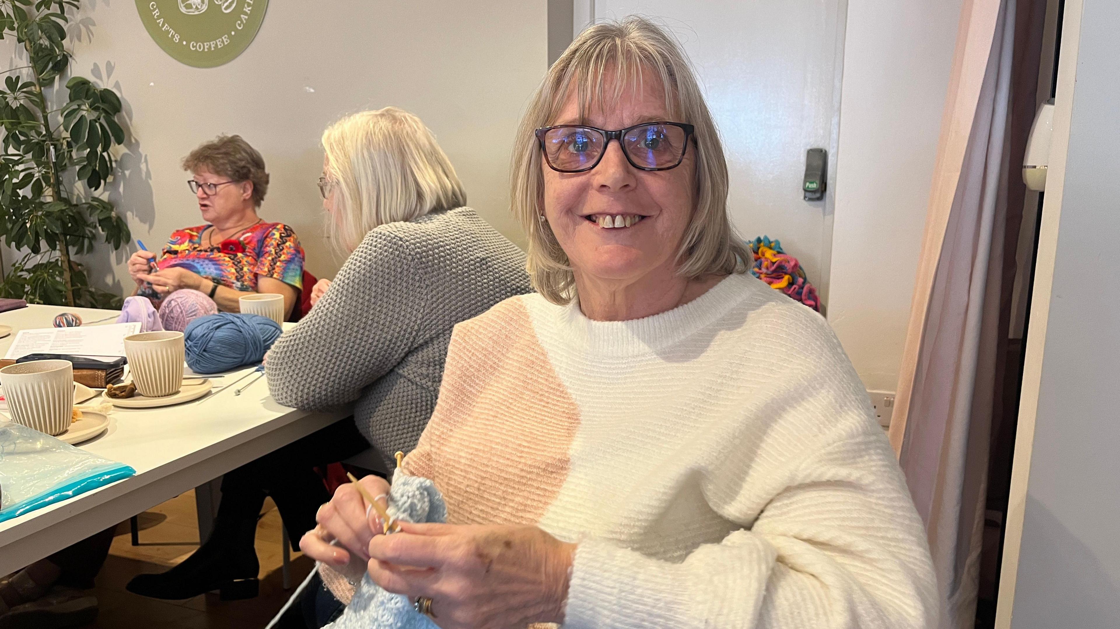 A woman with shoulder-length blonde hair and black glasses. She is wearing a white and pink jumper, and is holding blue knitting wool and knitting needles.
