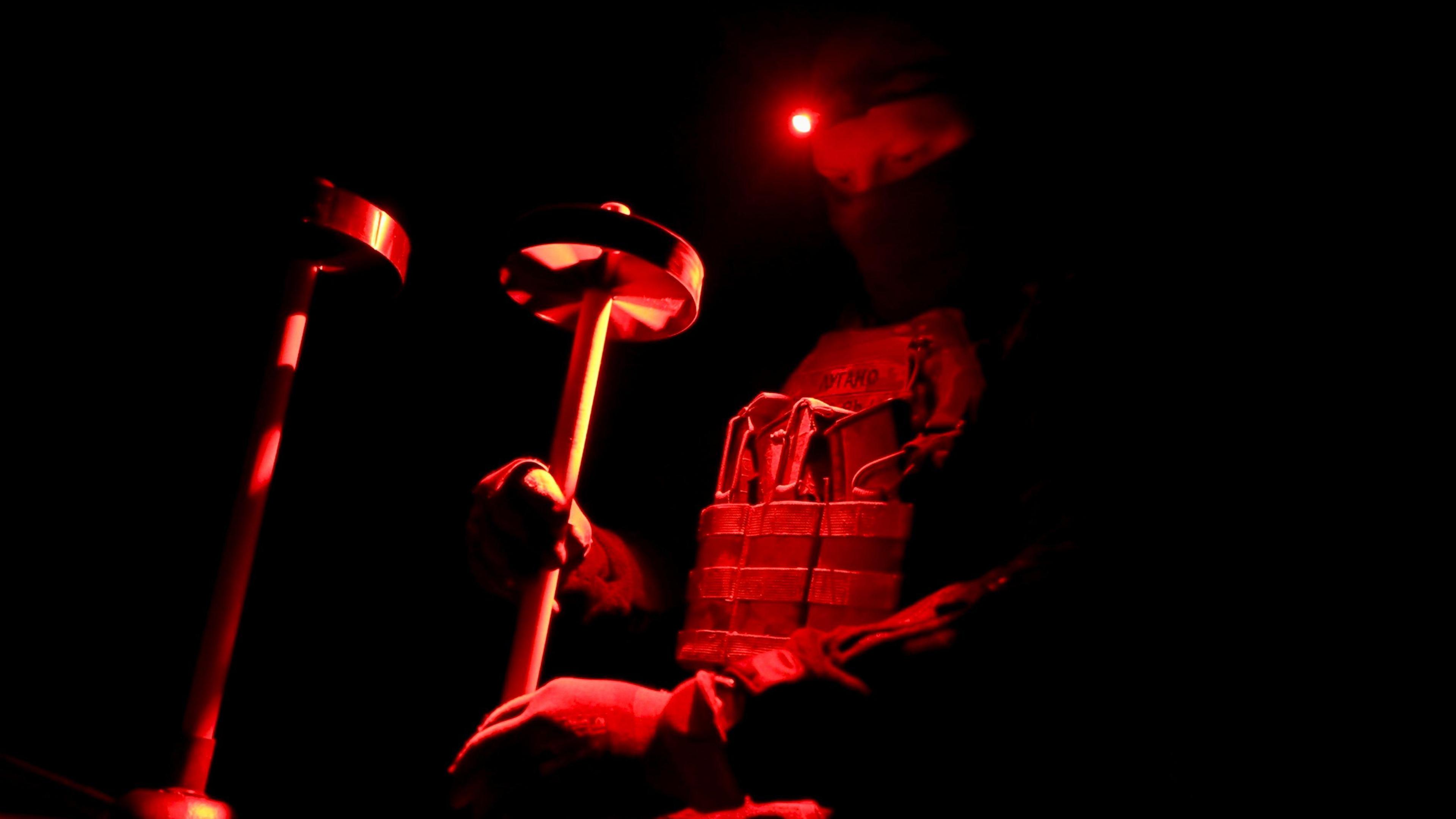 A man wearing a balaclava adjusts drone equipment in the darkness, under red light