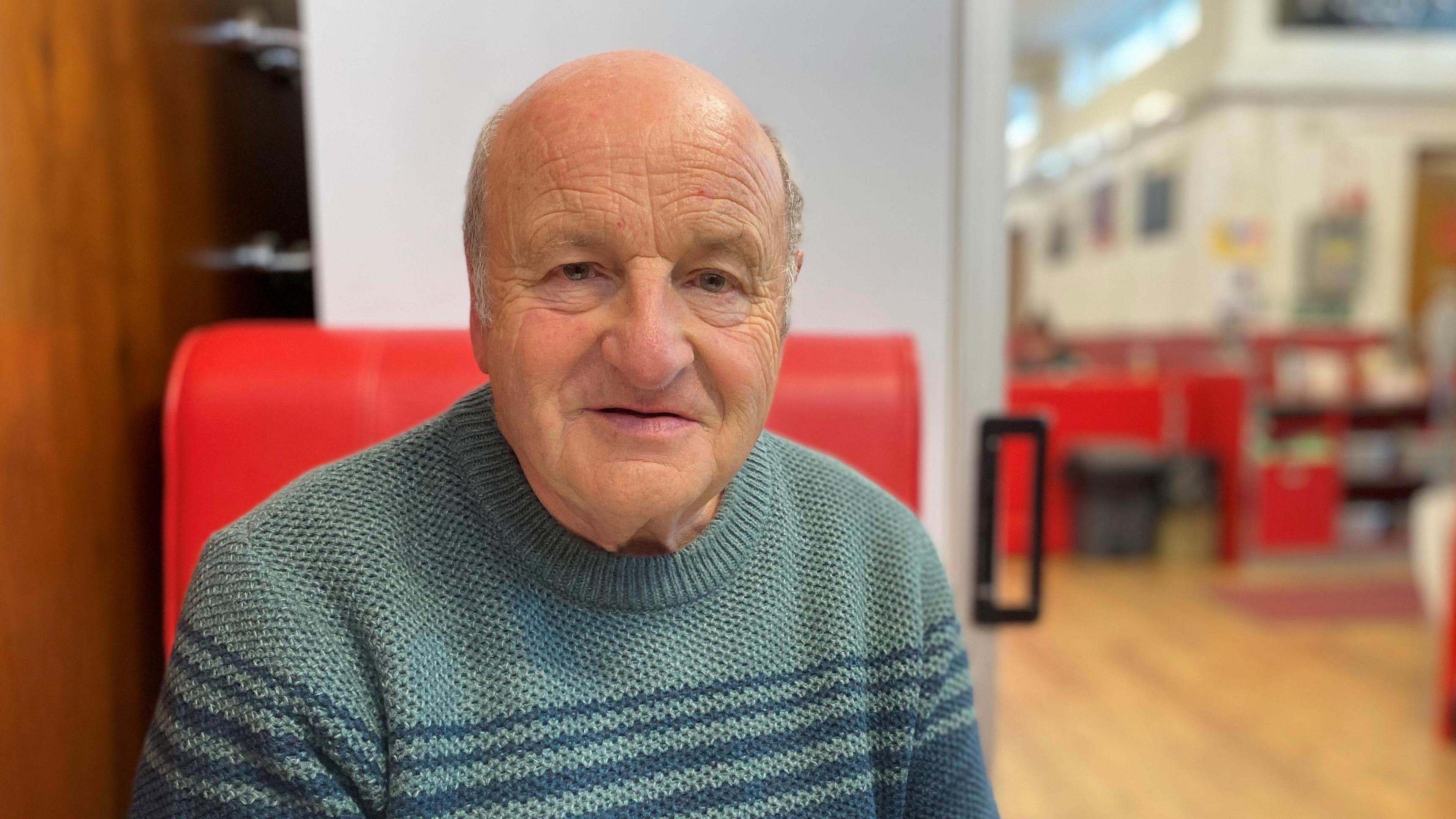 An old bald man in a blue striped jumper sits in a red chair and looks at the camera, slightly perplexed.